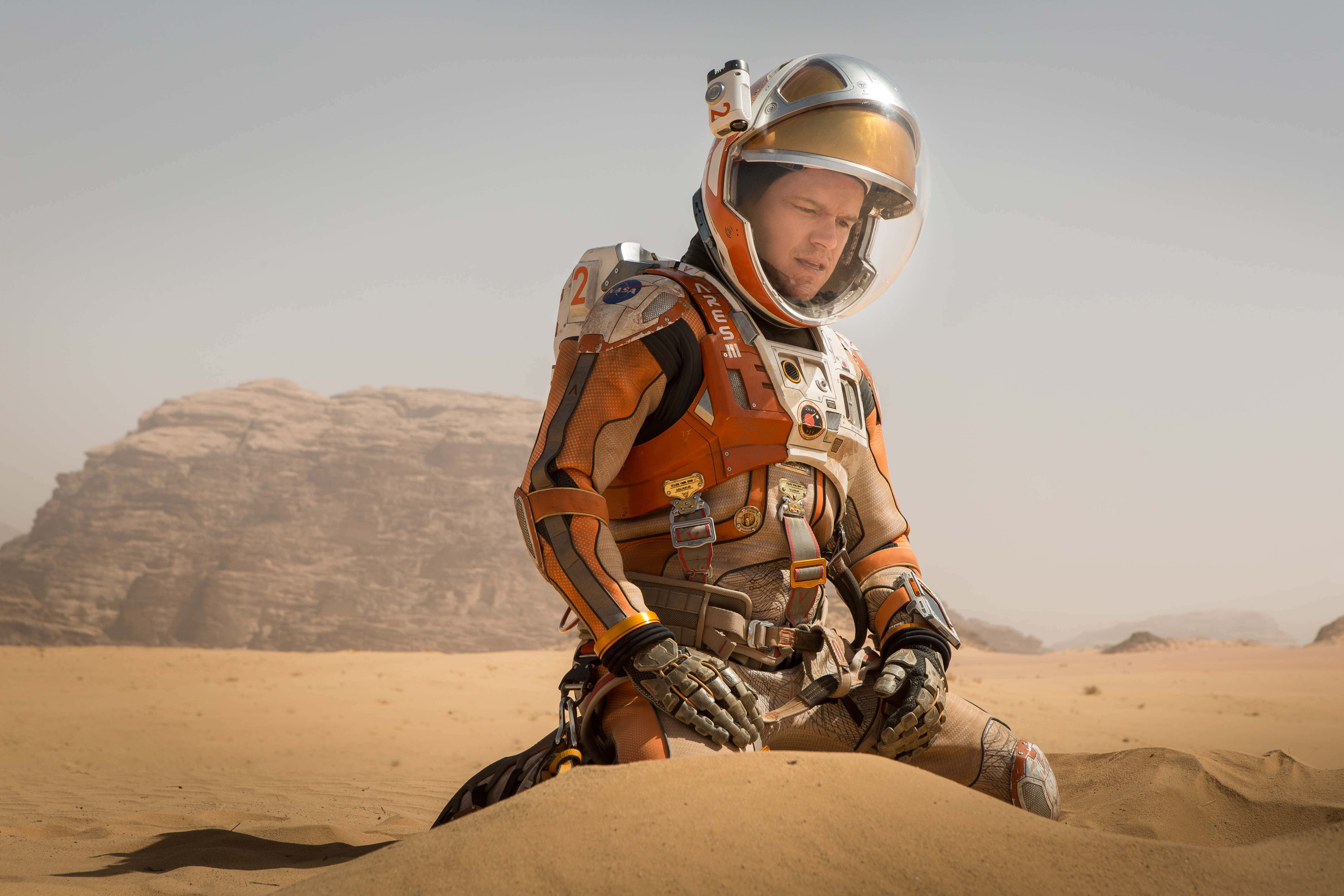 The Martian 桌布 壁紙 — 一名穿著橙色太空衣的太空人在朦朧天空下，跪在岩石丘陵遍佈的荒蕪沙漠中。