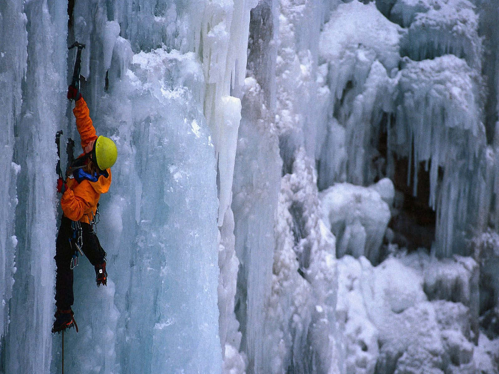 Ice Climbing 桌布 壁紙 — 一名穿著橙色裝備的攀冰者正在攀登一座高聳的冰凍瀑布，下方懸掛著冰柱。
