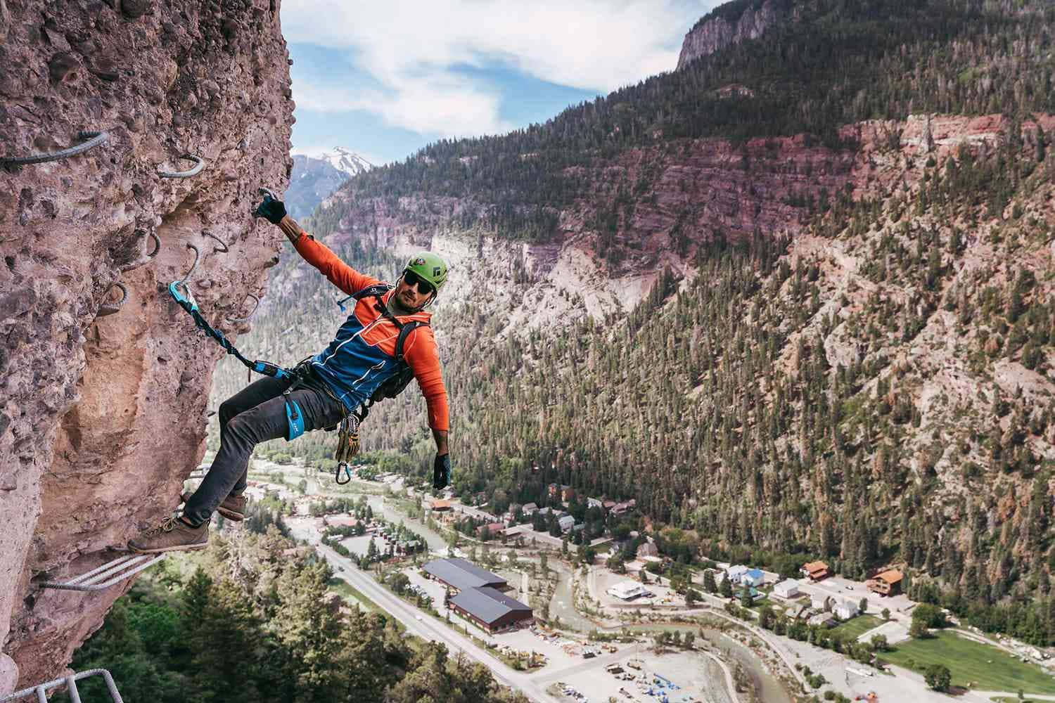 Colorado Mountain 桌布 壁紙 — 一名穿著橙色裝備的攀岩者正在攀登陡峭的懸崖，俯瞰著森林覆蓋的山谷和遠處白雪覆蓋的山峰，天空湛藍。