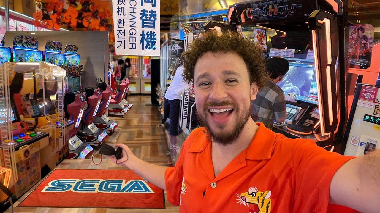 日本 桌布 壁紙 — 一名穿著橙色襯衫的微笑男子在一個光線明亮、充滿Sega遊戲機和發光螢幕的遊戲機中心自拍。