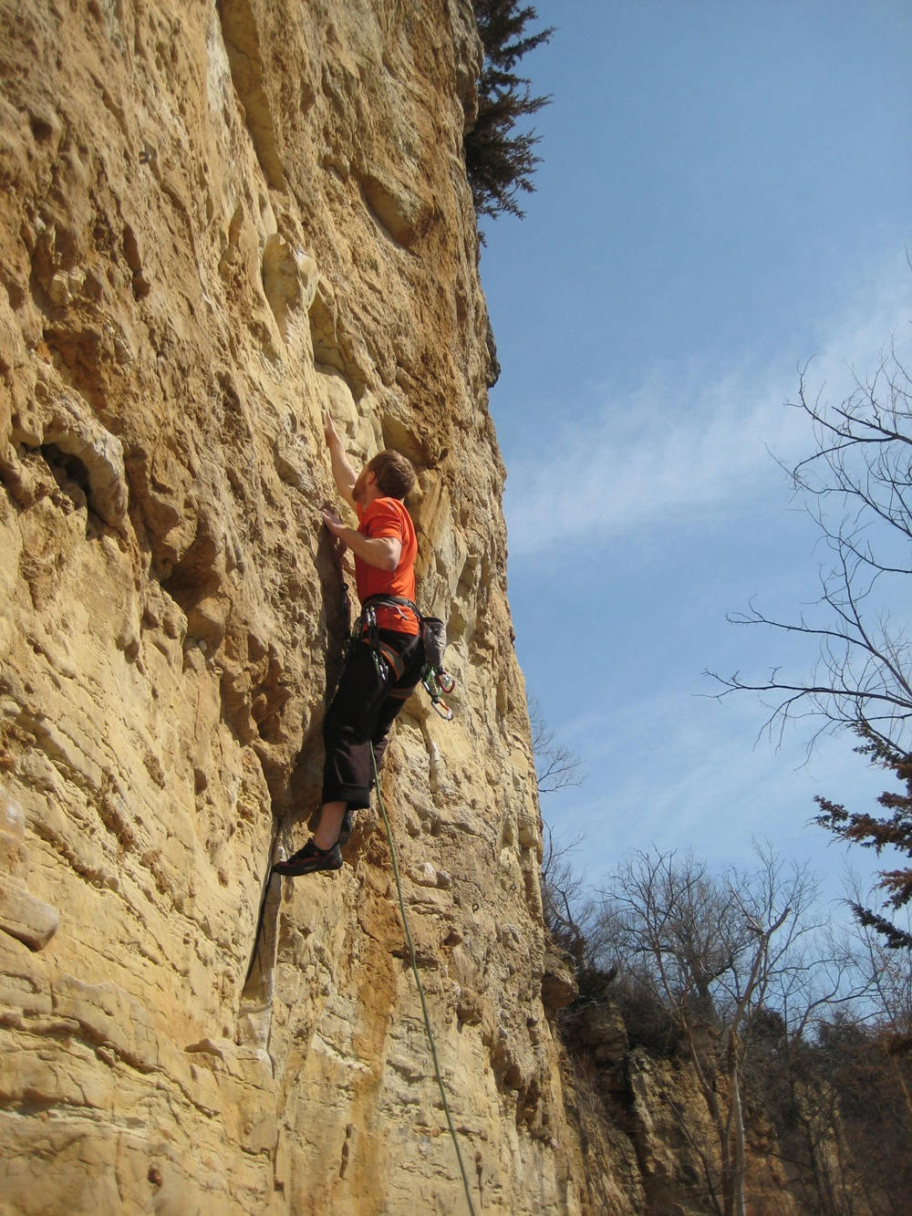 Climbing 桌布 壁紙 — 一名穿著橙色襯衫的攀岩者在晴朗的藍天下攀爬陽光照射的棕褐色懸崖面，下方有光禿禿的樹木。