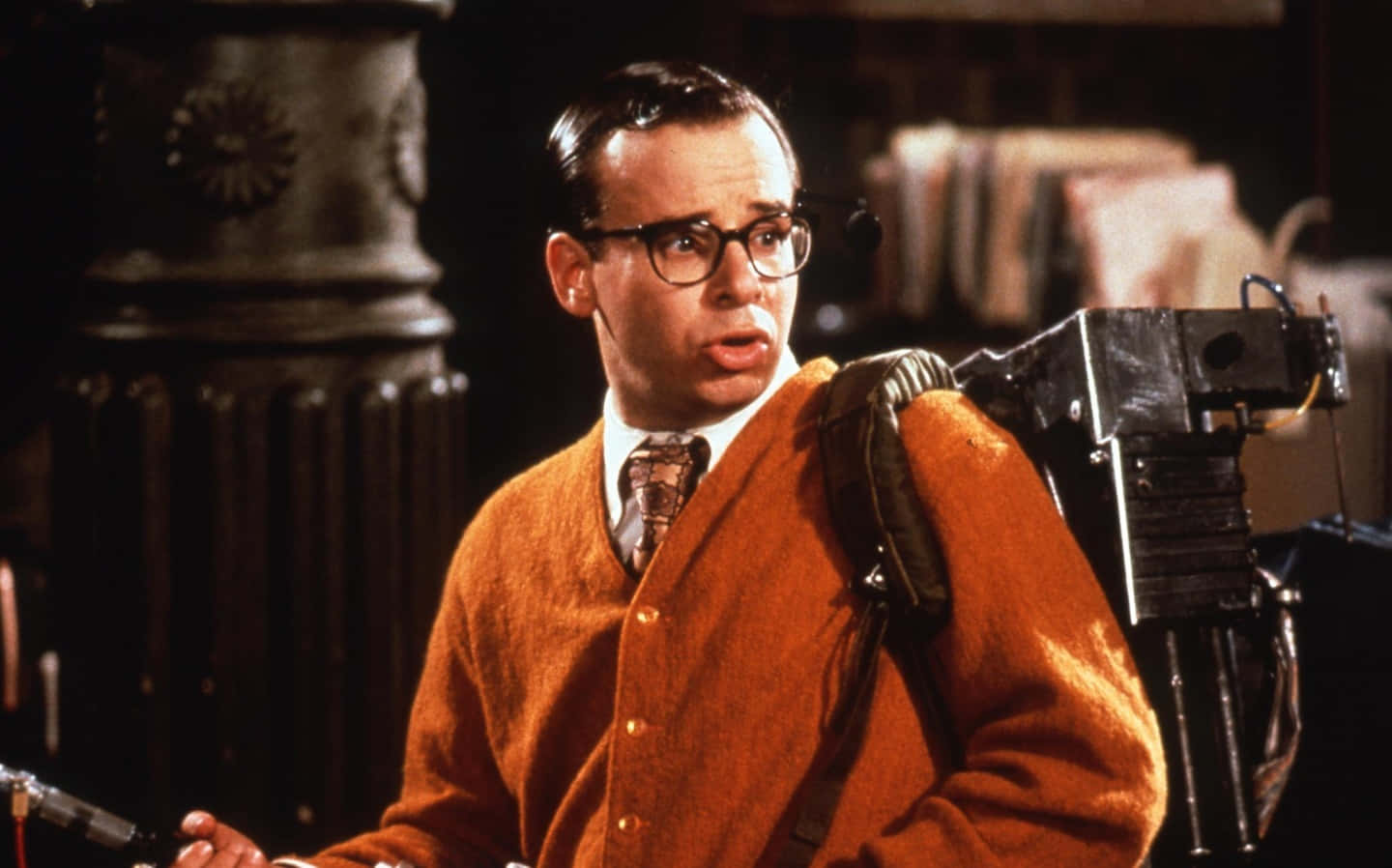 rick moranis 桌布 壁紙 — 一名穿著橙色開襟毛衣和眼鏡的男子，帶著驚訝的表情背著一個笨重的質子背包。