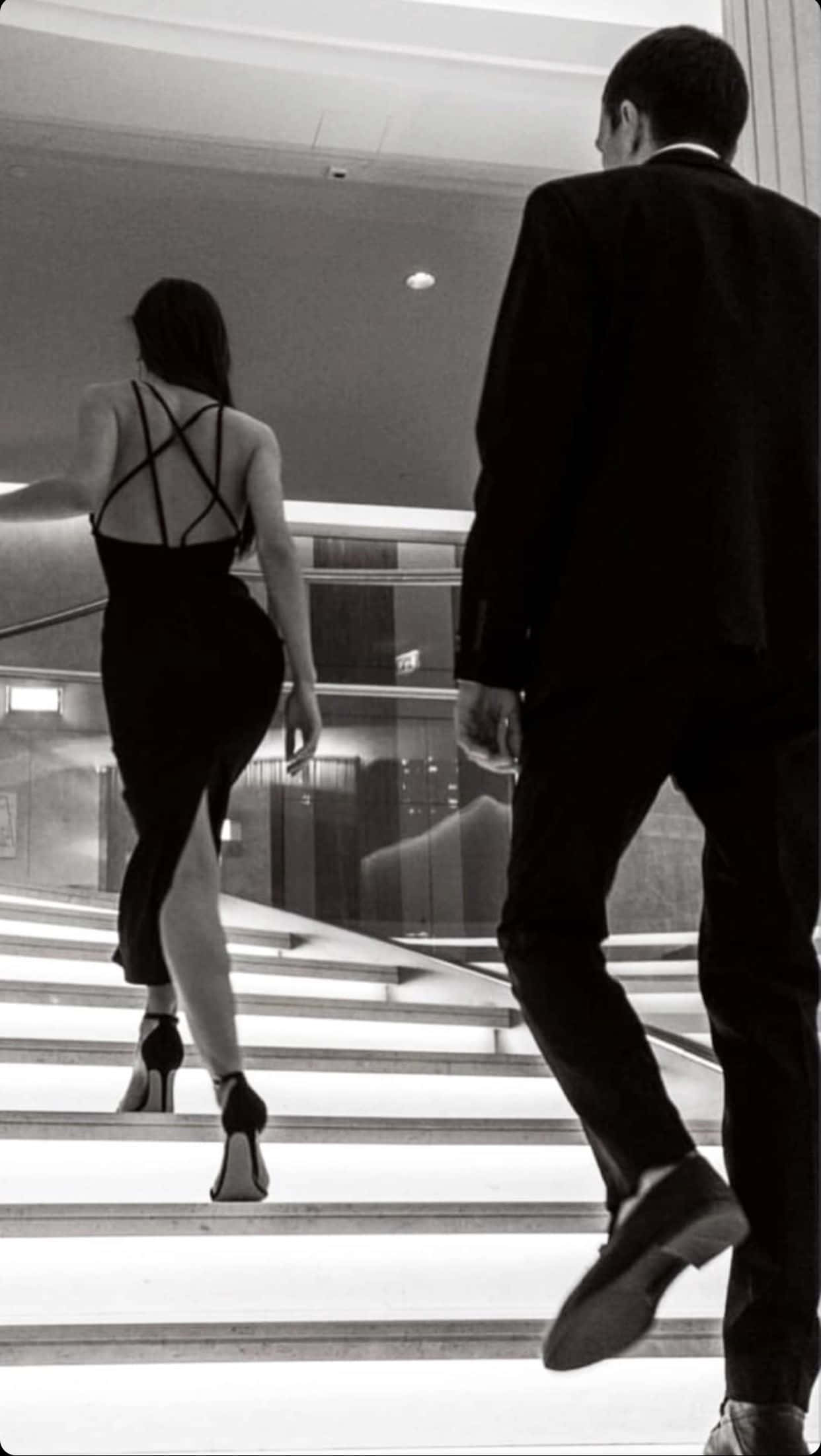 Aesthetic Couple 桌布 壁紙 — 一名穿著正式服裝的男士和女士在時尚的單色室內空間中登上現代樓梯。