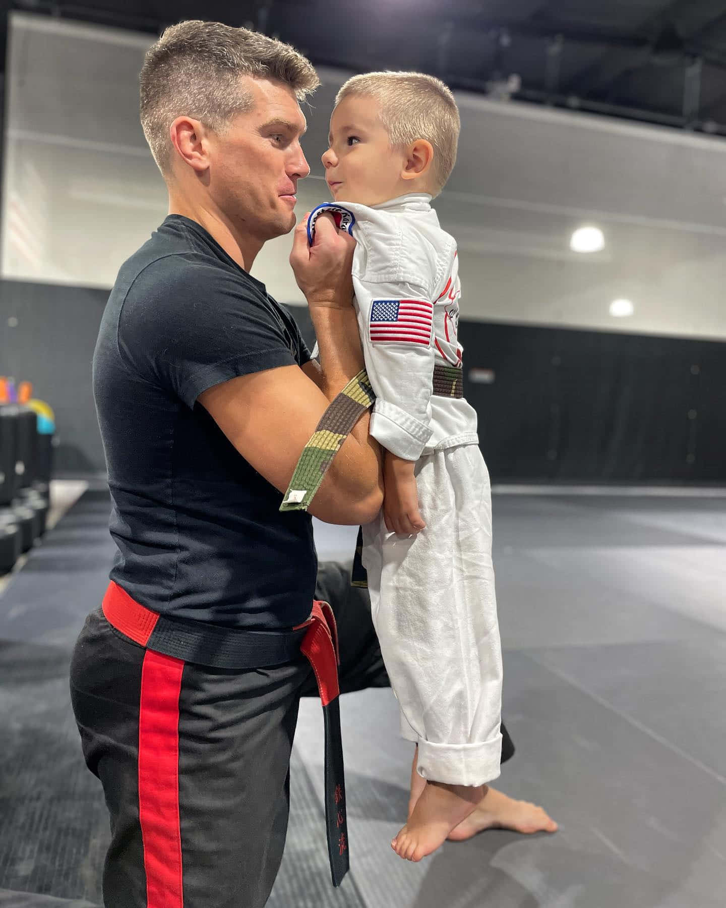 MMA 桌布 壁紙 — 一名穿著武術裝備的男子在健身房內抱著一個穿著白色道服、帶有美國國旗補丁的小孩。