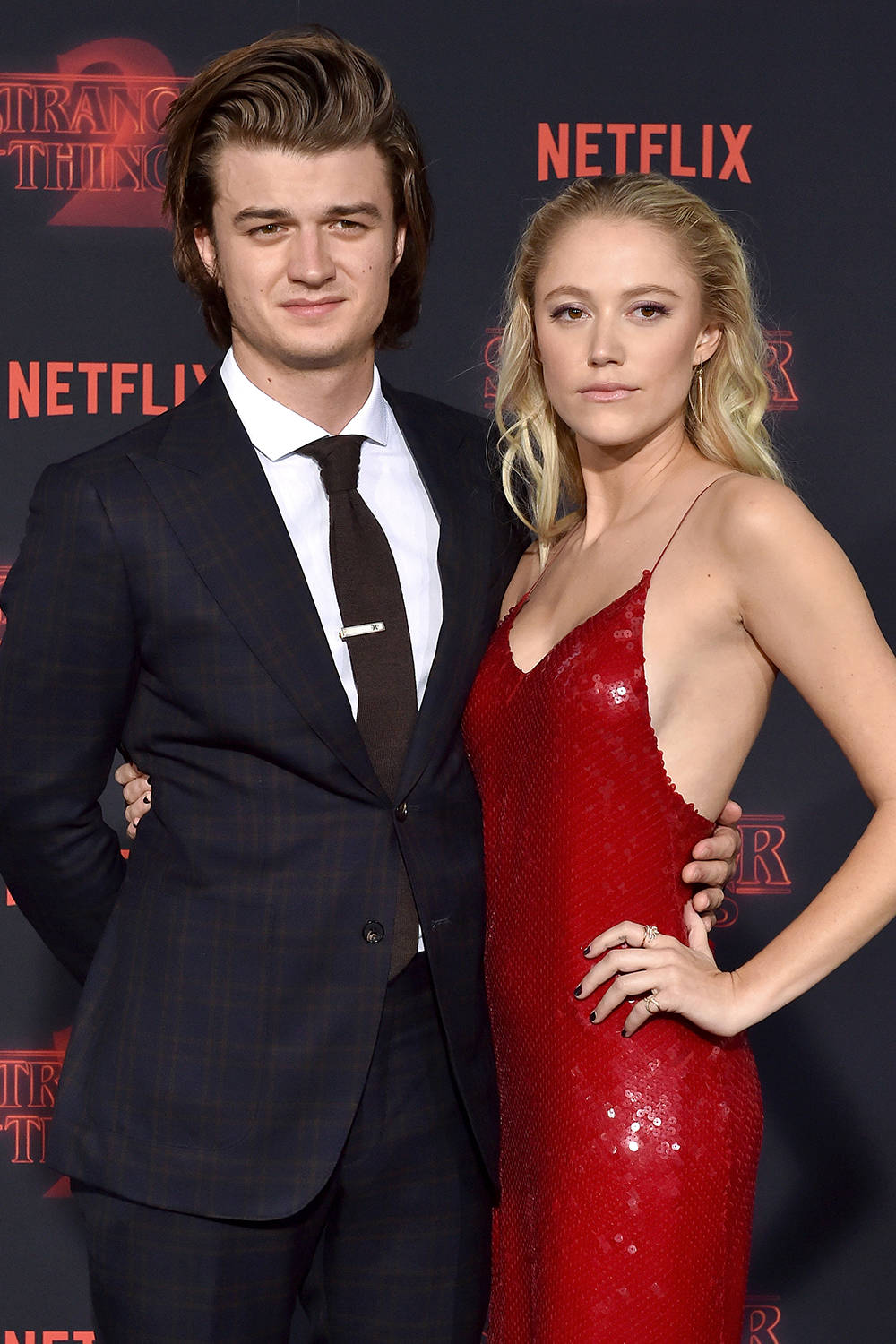 Joe Keery 桌布 壁紙 — 一名穿著深色格紋西裝的男子和一名穿著閃亮紅色連衣裙的女子在 Netflix 活動中，以《怪奇物語2》背景一起擺姿勢。