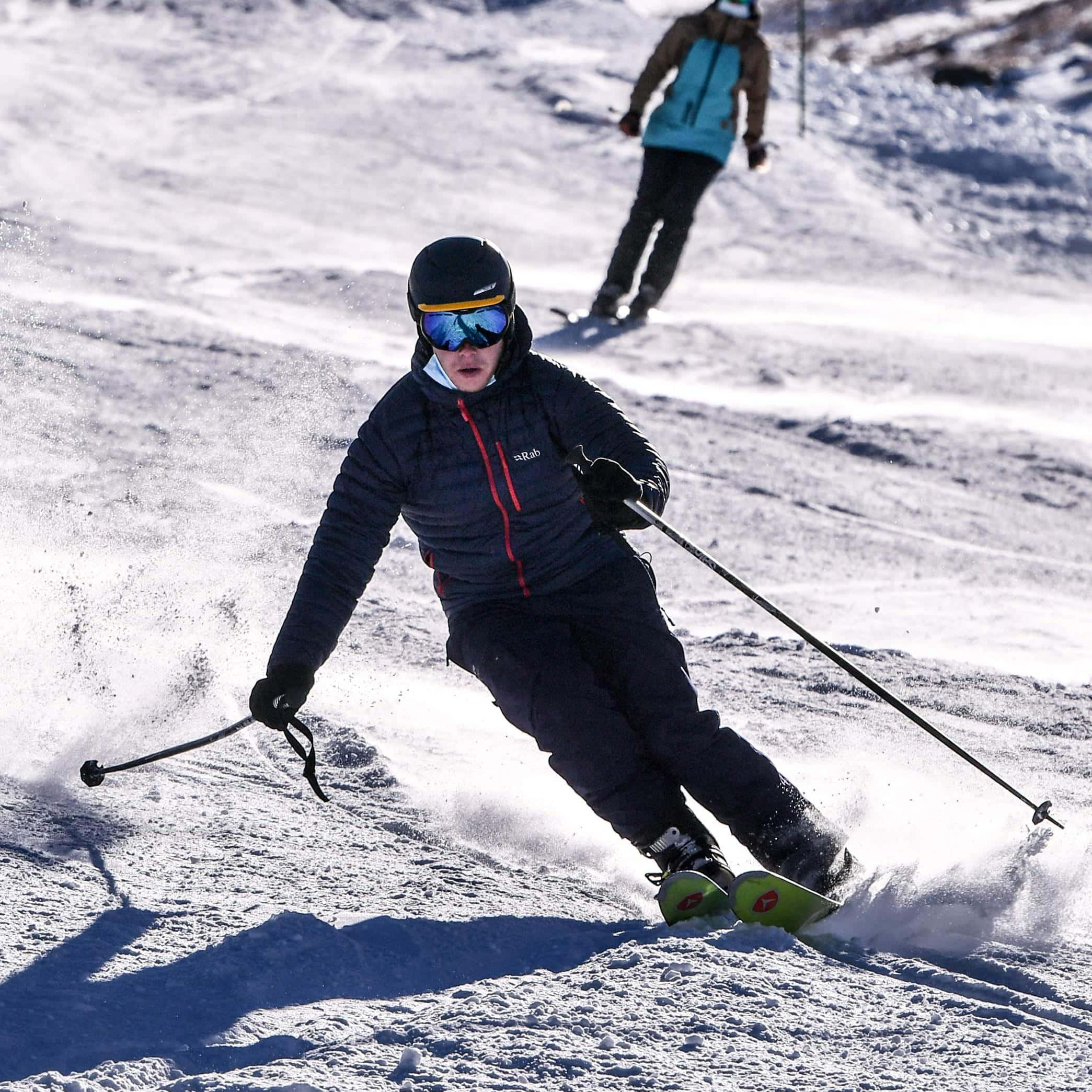 ski 桌布 壁紙 — 一名穿著深色裝備的滑雪者在陽光明媚的雪坡上穿過新雪，激起粉雪飛揚。