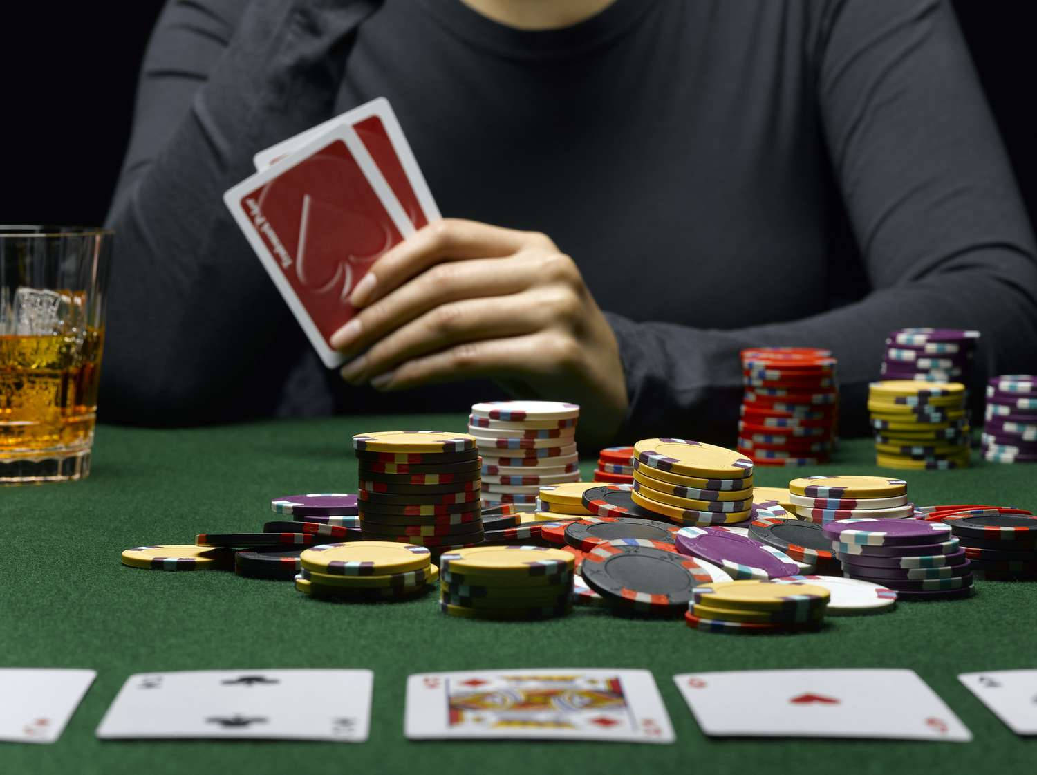 Playing Cards 桌布 壁紙 — 一名穿著深色襯衫的人，在一個散落著彩色籌碼和面朝上紙牌的綠色撲克桌上，手持兩張紅色背面的撲克牌。