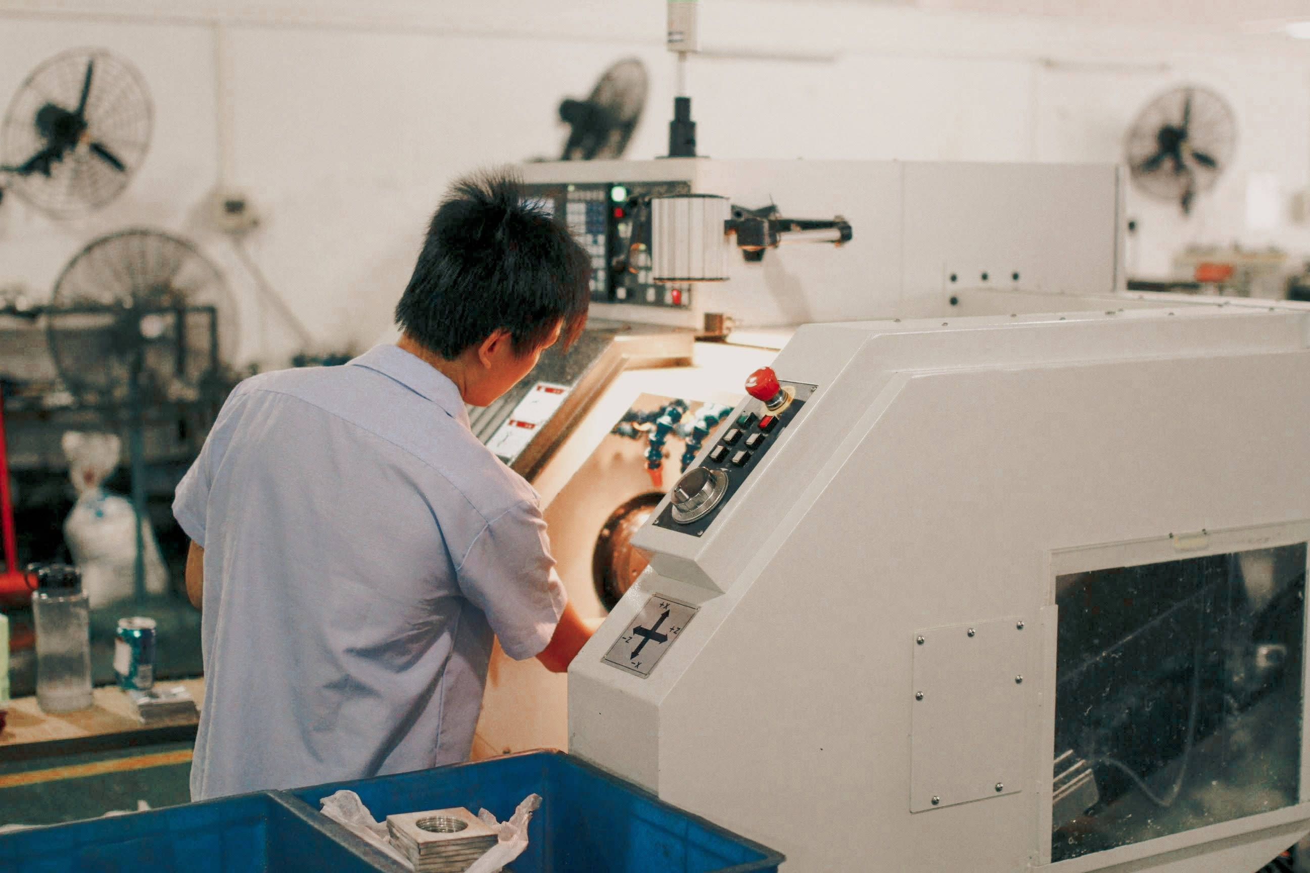 machines 桌布 壁紙 — 一名穿著淺藍色襯衫的工人在工廠內操作一部大型白色工業機器，周圍有吊扇和工具。
