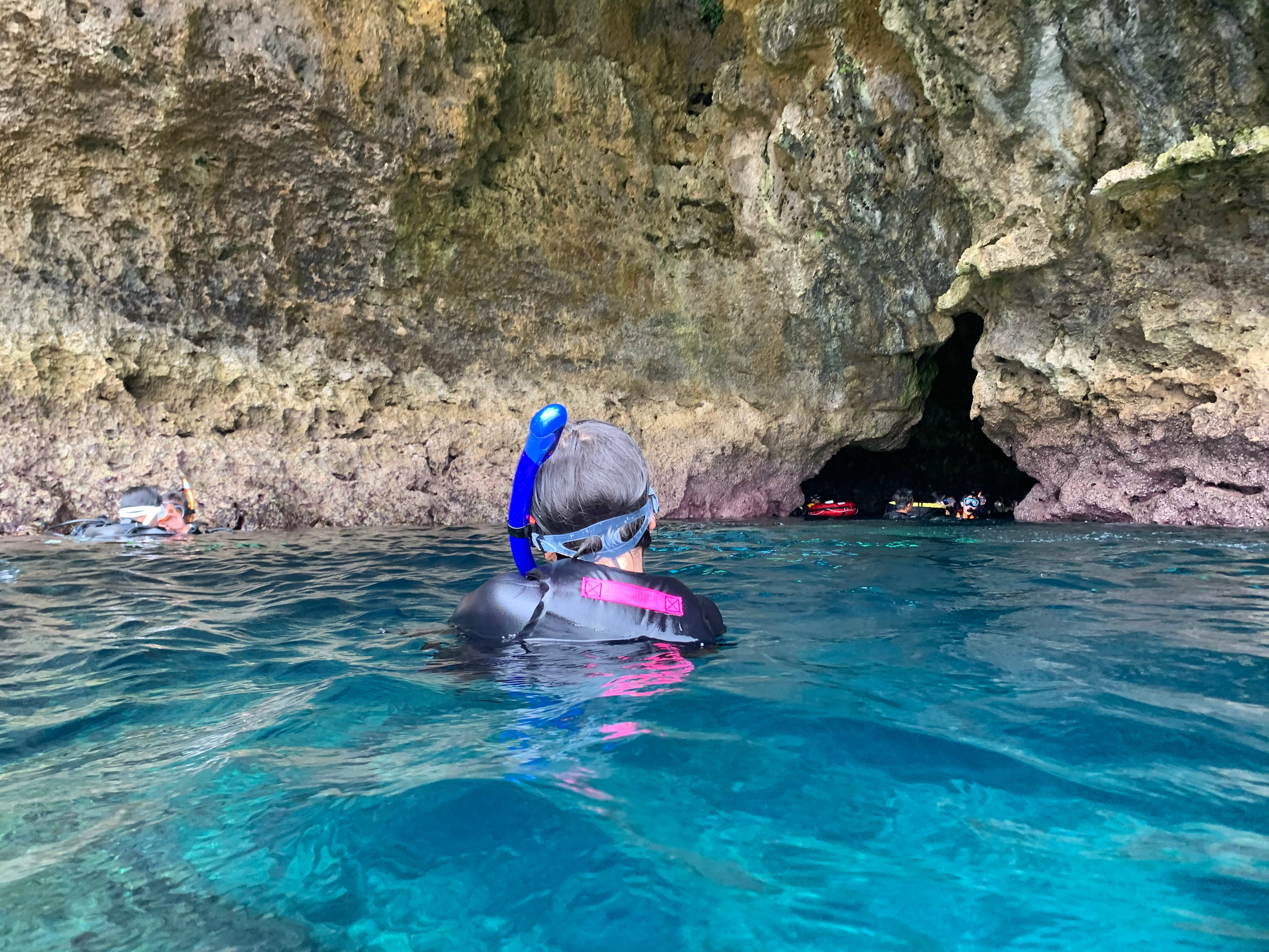 Snorkeling 桌布 壁紙 — 一名穿著潛水衣的浮潛者從清澈的碧綠海水中凝視著一個被崎嶇懸崖環繞的黑暗海蝕洞。