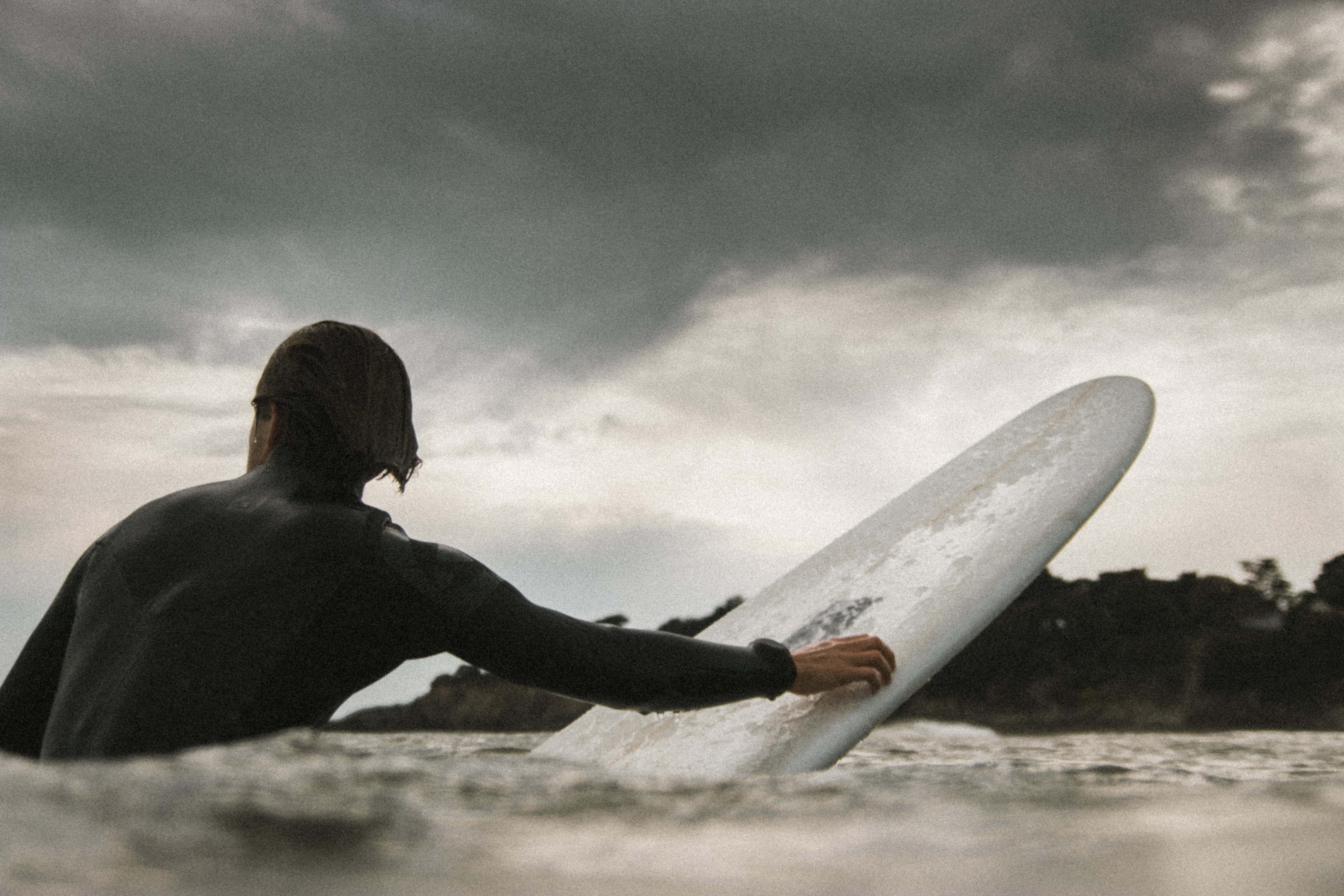 surf 桌布 壁紙 — 一名穿著潛水衣的衝浪者抓住衝浪板，在風暴般的灰色天空下划過波濤洶湧的海水。
