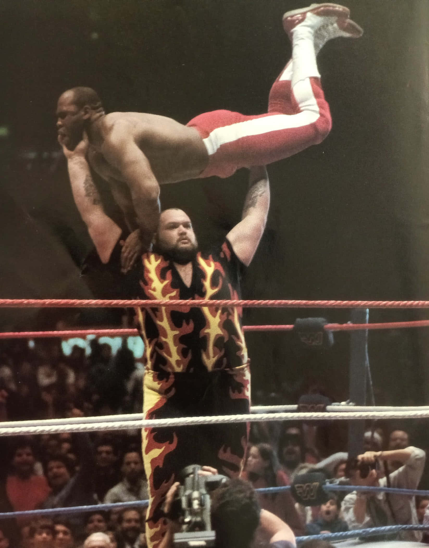 WWE 桌布 壁紙 — 一名穿著火焰圖案短褲的摔角手在明亮的場館燈光下，在摔角台上將另一名摔角手高舉過頭。