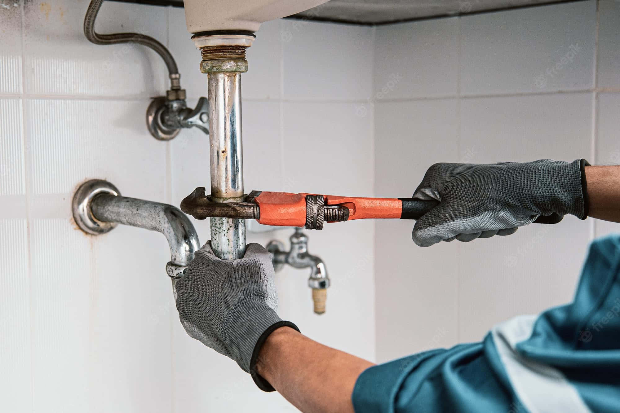 Plumbing 桌布 壁紙 — 一名穿著灰色手套的水管工，用橙色扳手在磁磚牆下的白色洗手台下方鎖緊一條生鏽的水管。