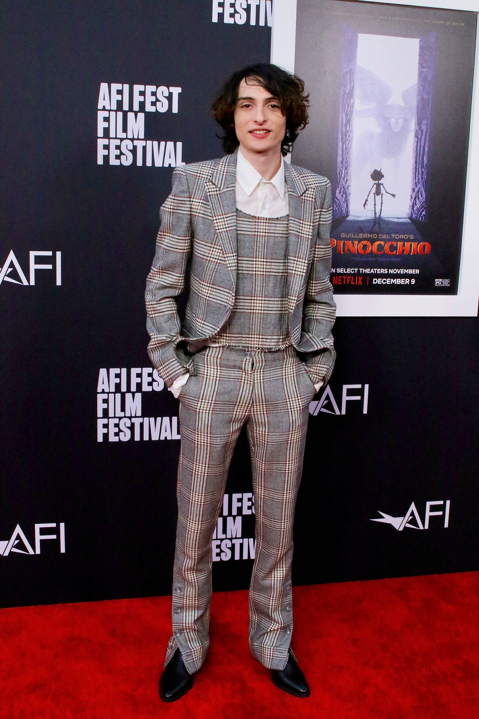 finn wolfhard 桌布 壁紙 — 一名穿著灰色格紋西裝的男子自信地站在AFI Fest電影節的紅地毯上，身後有一張皮諾丘電影海報。