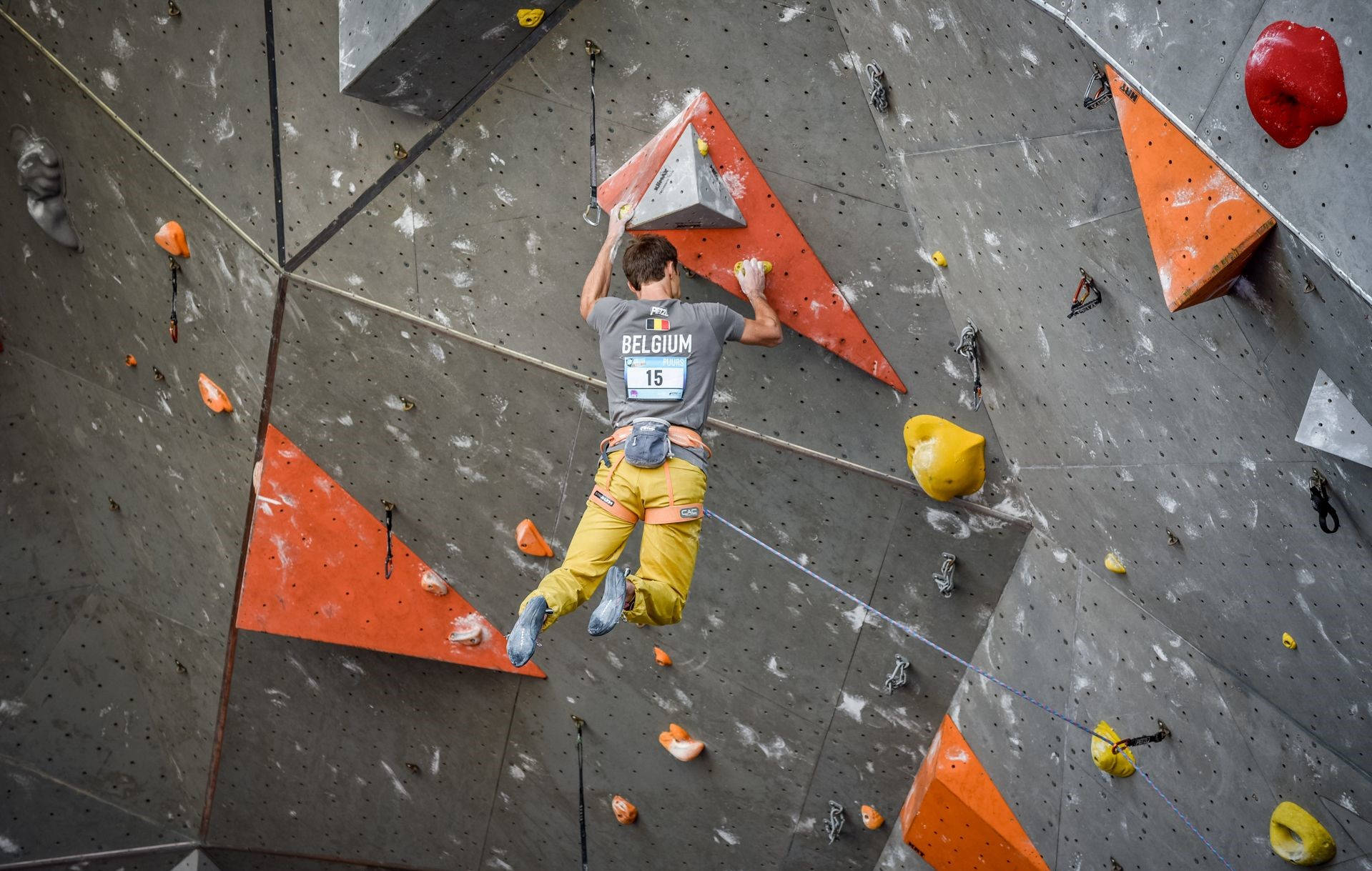 Climbing 桌布 壁紙 — 一名穿著灰色比利時球衣和黃色褲子的攀岩者，攀爬帶有橙色和黃色握點的紋理室內攀岩牆。