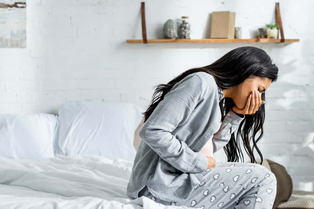Pregnant 桌布 壁紙 — 一名穿著灰色睡衣的女人坐在床上，雙手抱頭，在明亮的白色臥室中看起來很苦惱。