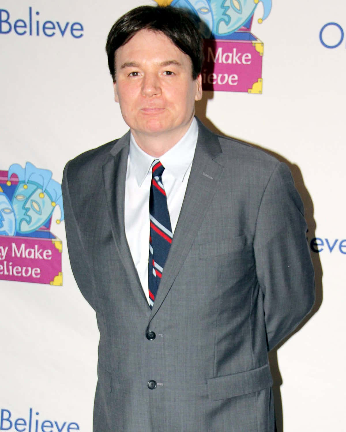 Mike Myers 桌布 壁紙 — 一名穿著灰色西裝和條紋領帶的男子自信地站在一個『Make Believe』活動中，身後有彩色標誌。