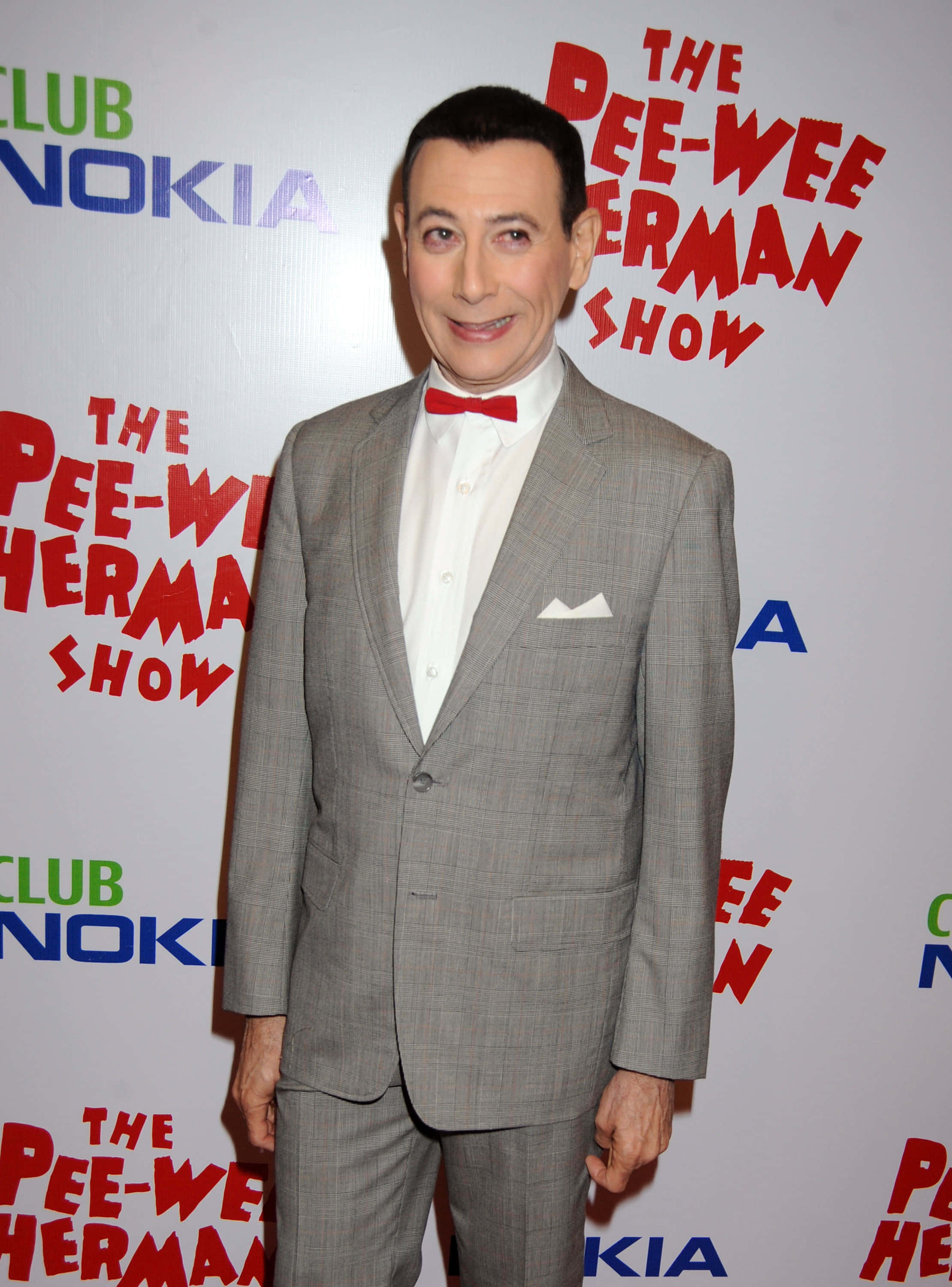 Paul Reubens 桌布 壁紙 — 一名穿著灰色西裝和紅色領結的男子在The Pee-wee Herman Show的背景前對著鏡頭微笑。