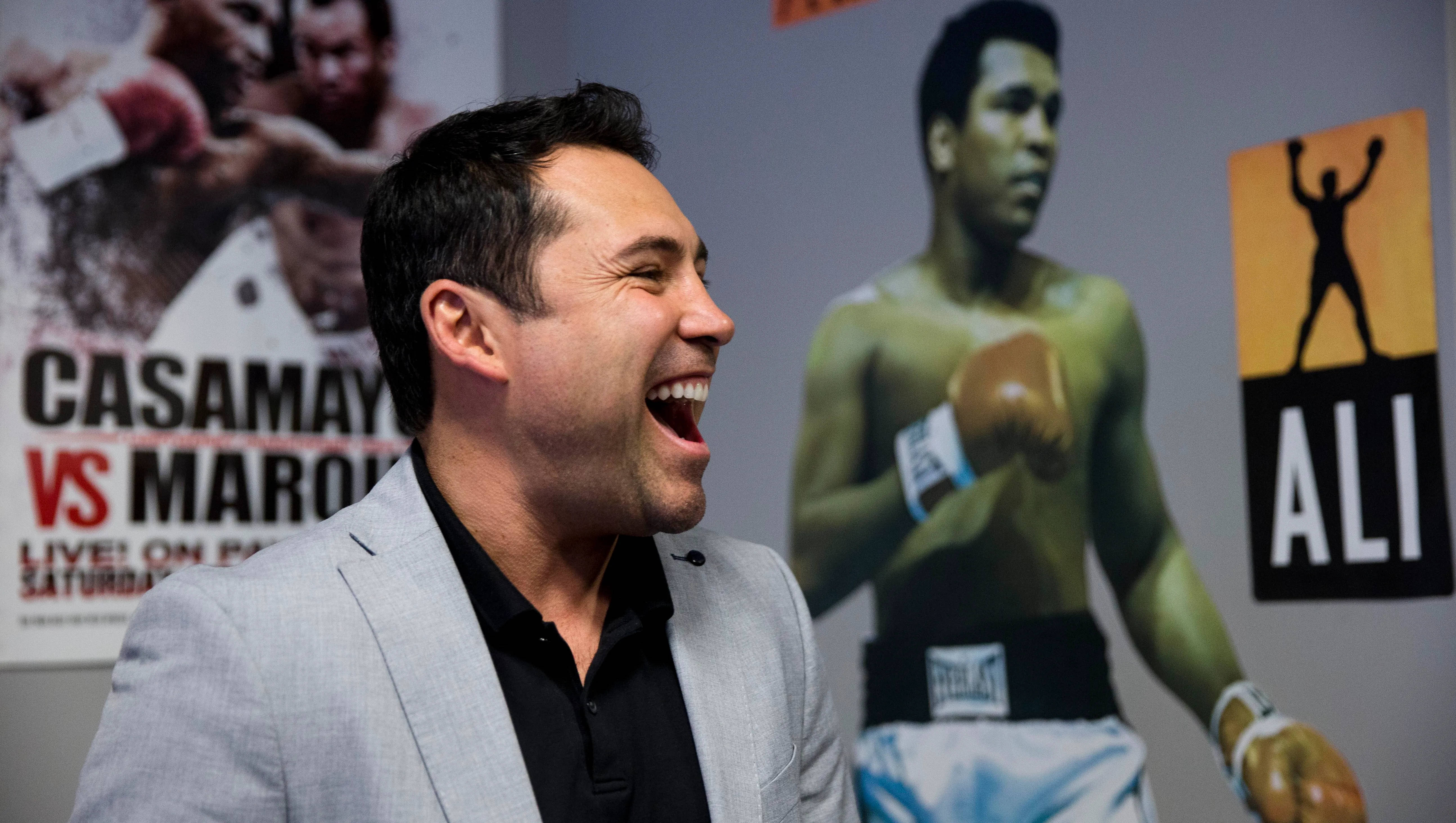 Boxing 桌布 壁紙 — 一名穿著灰色西裝的男子在拳擊活動中開懷大笑，身後有穆罕默德·阿里的海報。