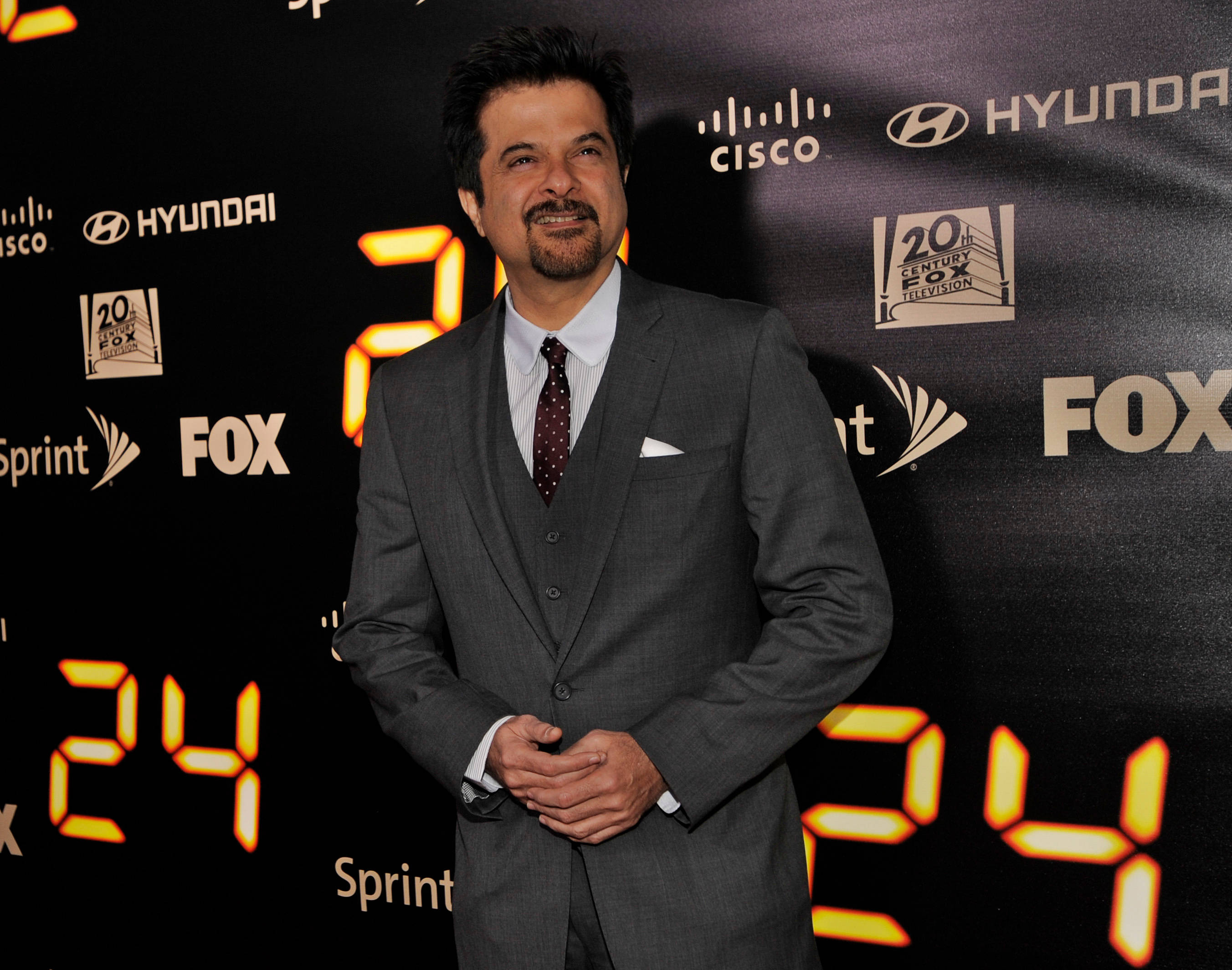 anil kapoor 桌布 壁紙 — 一名穿著灰色西裝的男子自信地站在黑色背景前，背景上有發光的橙色「24」數字和標誌。