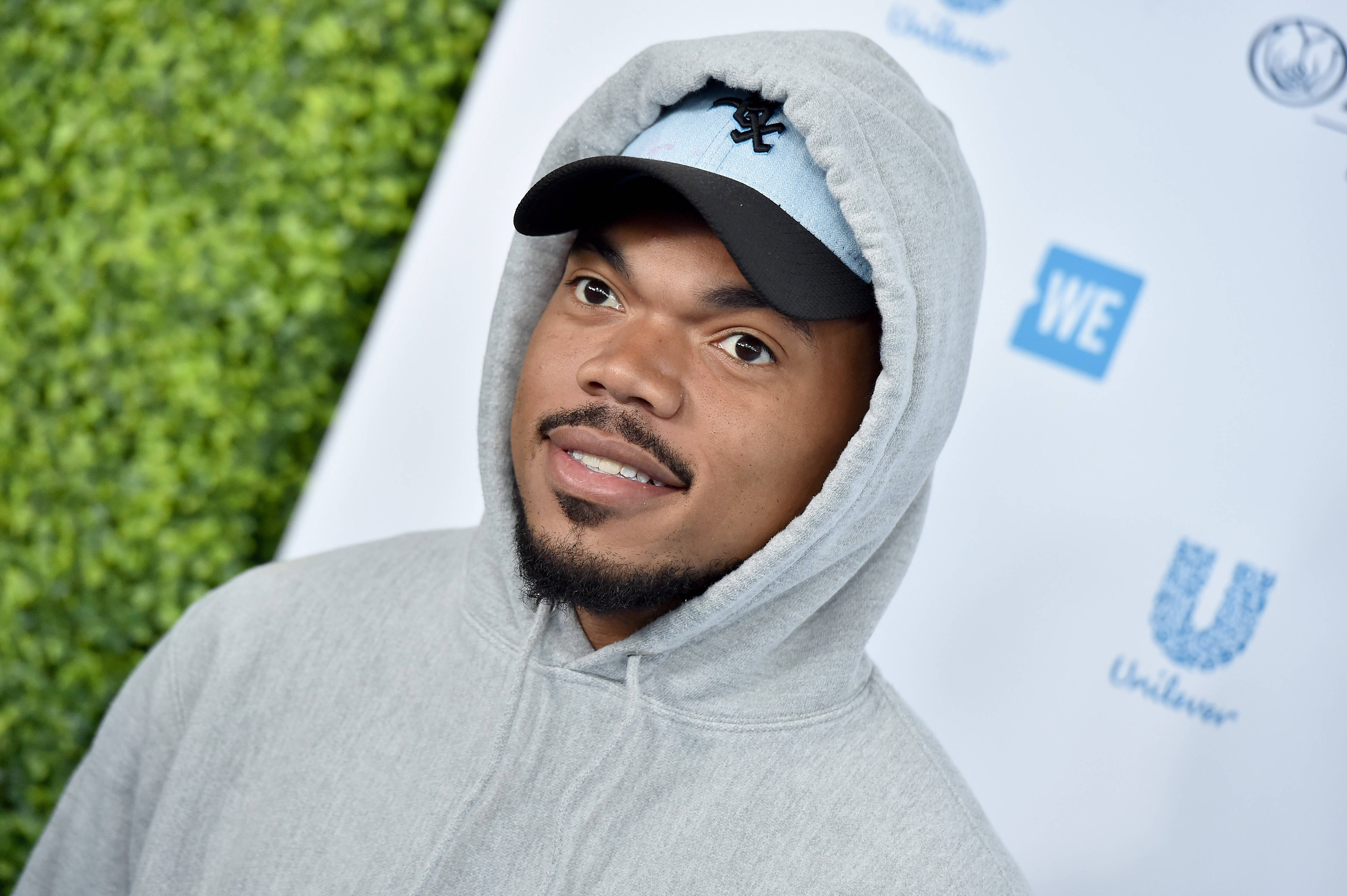 chance 桌布 壁紙 — 一名穿著灰色連帽衫和帽子的男子在綠色樹籬和品牌背景前對著鏡頭微笑。