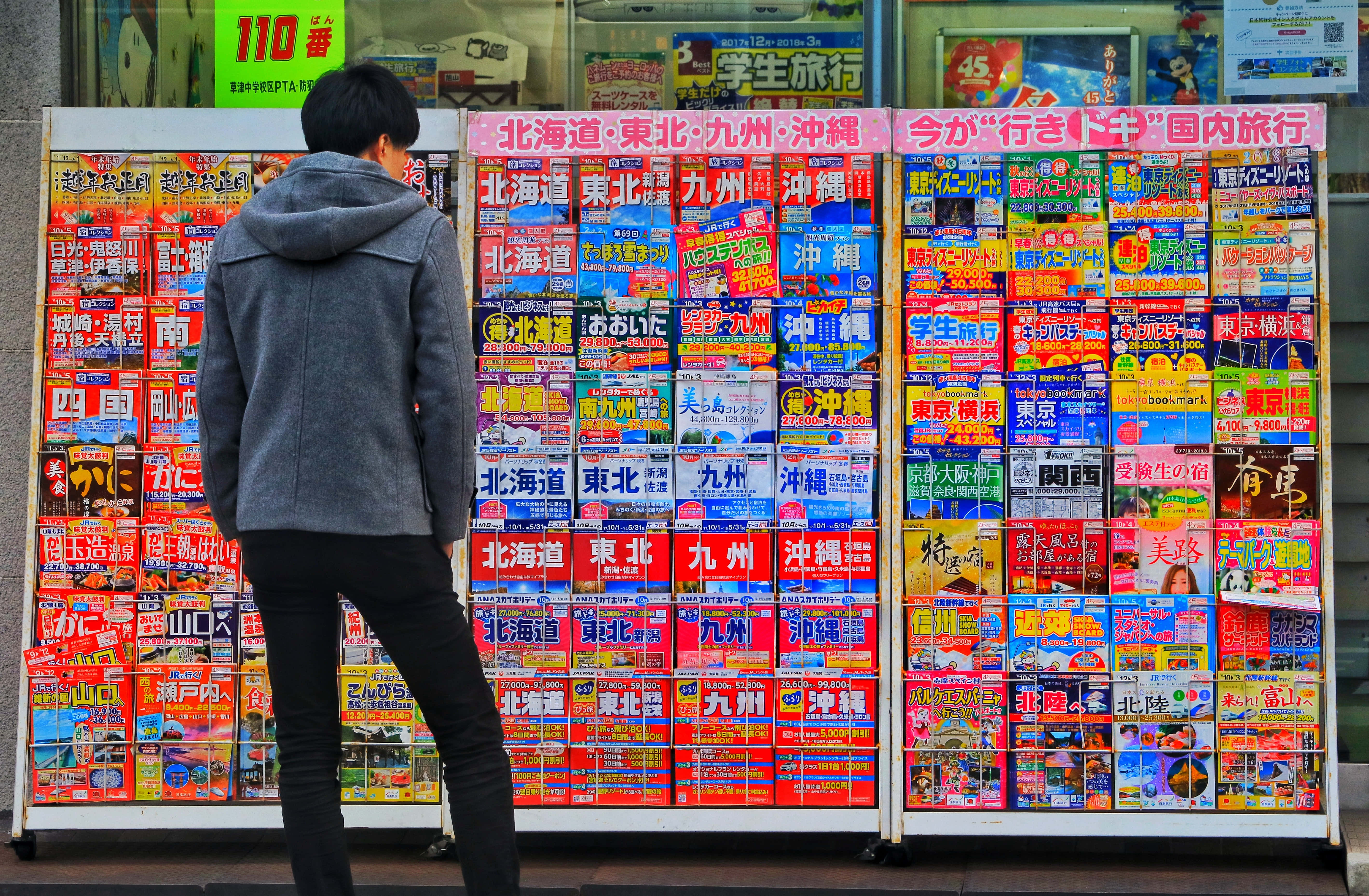 Store 桌布 壁紙 — 一名穿著灰色連帽衫的人站在城市商店櫥窗前，面前是一面色彩繽紛的日本旅遊手冊牆。