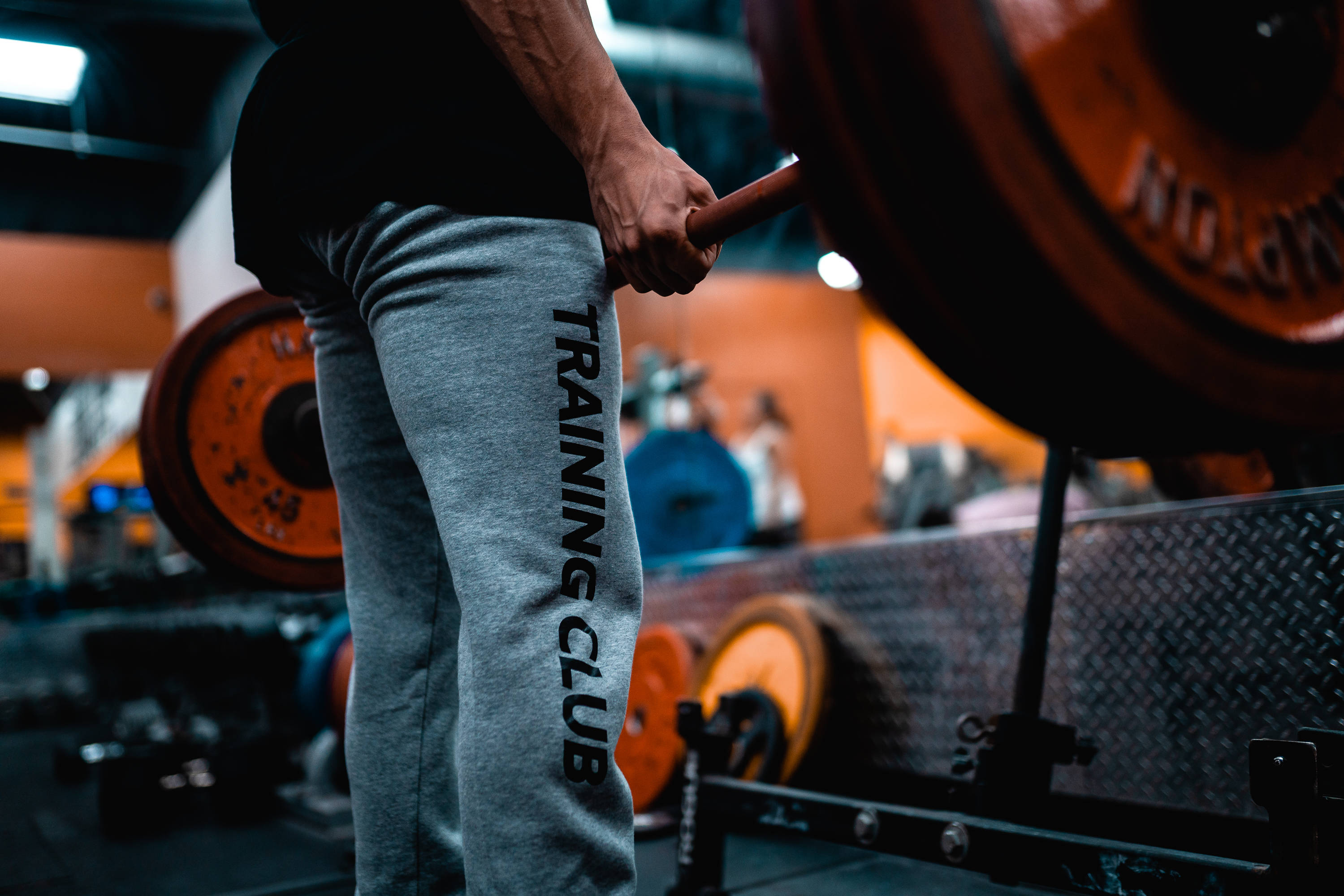 Weightlifting 桌布 壁紙 — 一名穿著灰色運動褲的人，在溫暖燈光的健身房中握住裝有橙色槓片的槓鈴。