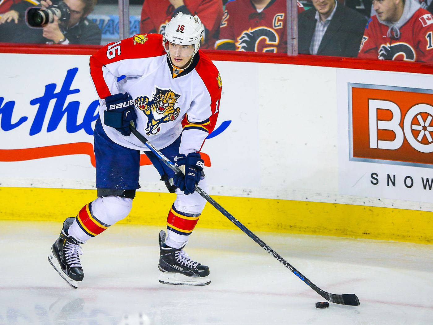 運動 桌布 壁紙 — 一名穿著白色、紅色和藍色球衣的佛羅里達美洲豹冰球球員，在比賽中帶著冰球在冰上滑行，背景有球迷和廣告。