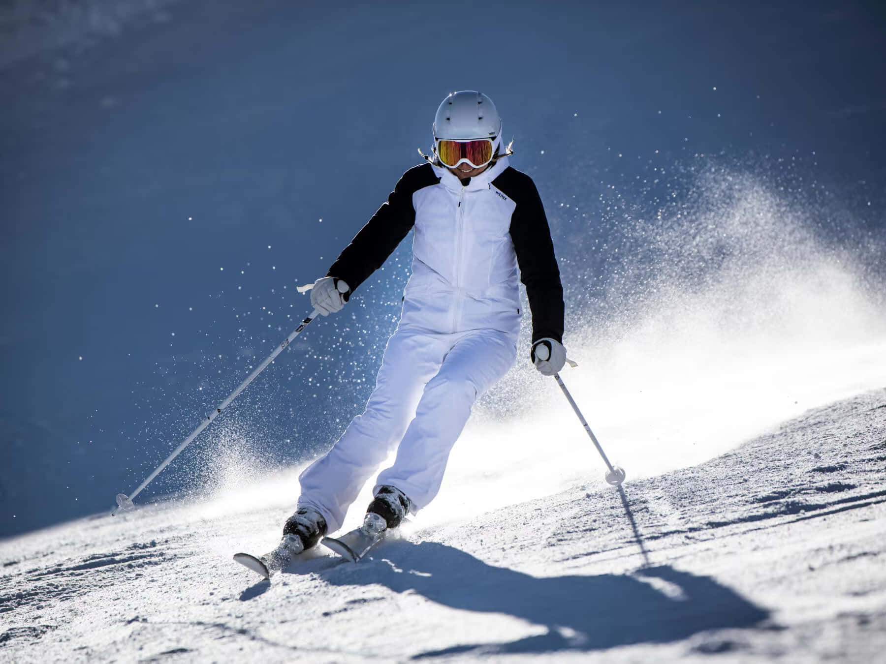 ski 桌布 壁紙 — 一名穿著白色裝備的滑雪者在雪坡上轉彎，將雪濺向明亮的藍天。