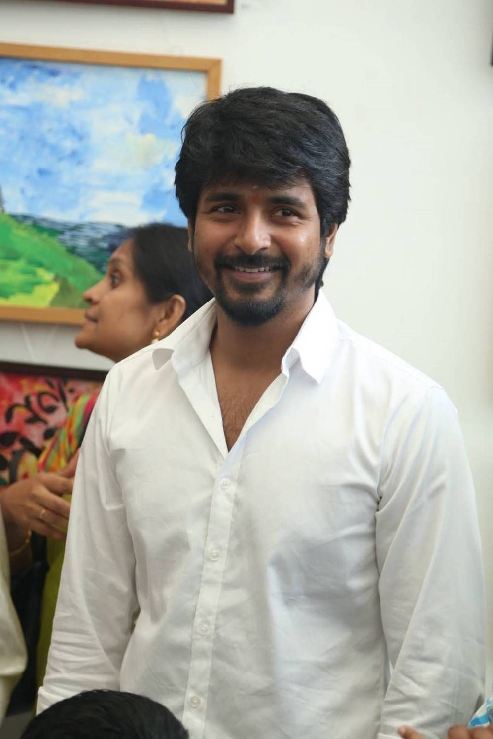 Sivakarthikeyan 桌布 壁紙 — 一名穿著白色襯衫的微笑男子站在色彩繽紛的藝術品前，身後隱約可見一名女子。