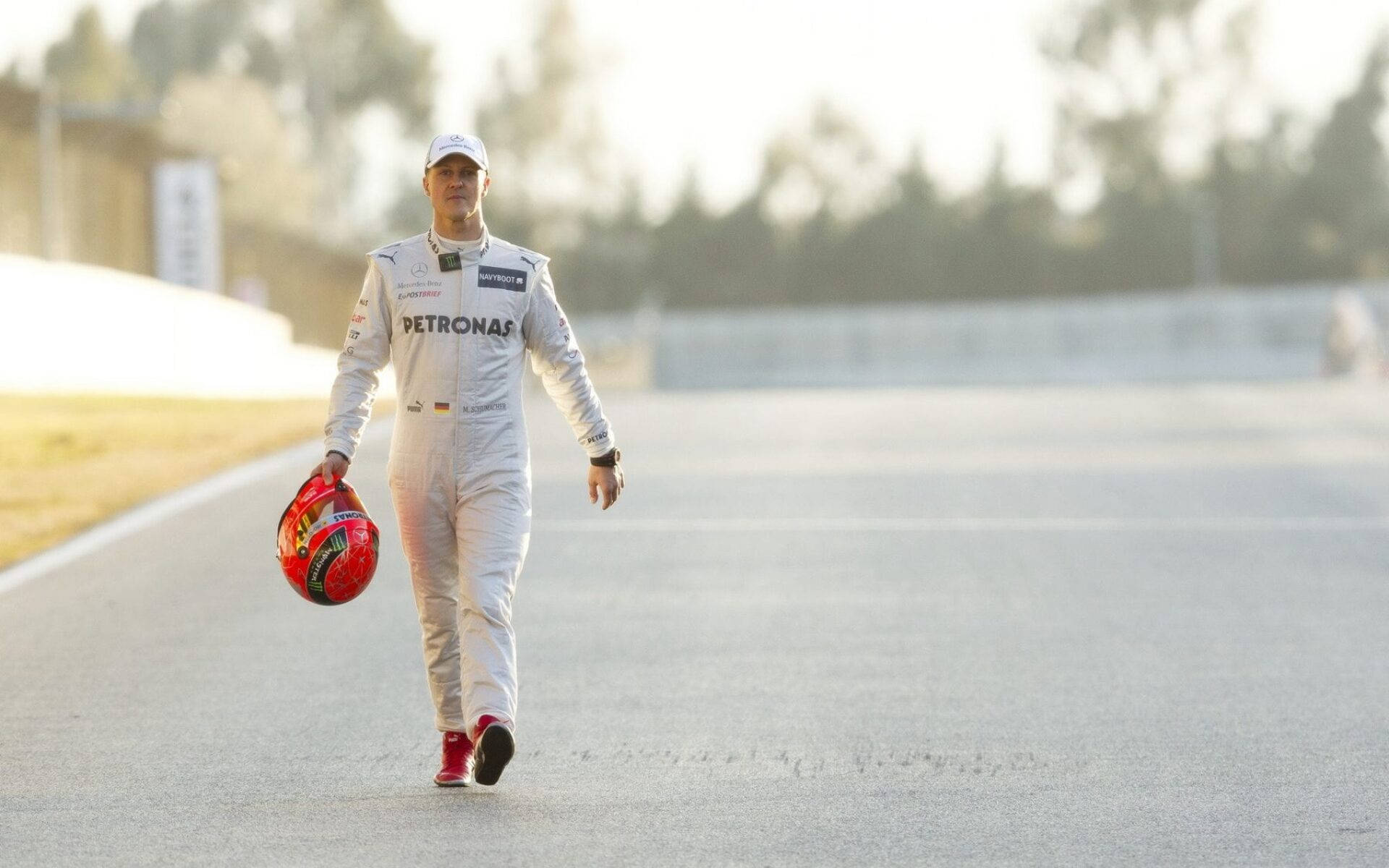 michael 桌布 壁紙 — 一名穿著白色賽車服的F1車手在日出時自信地走在空曠的賽道上，手持紅色頭盔。