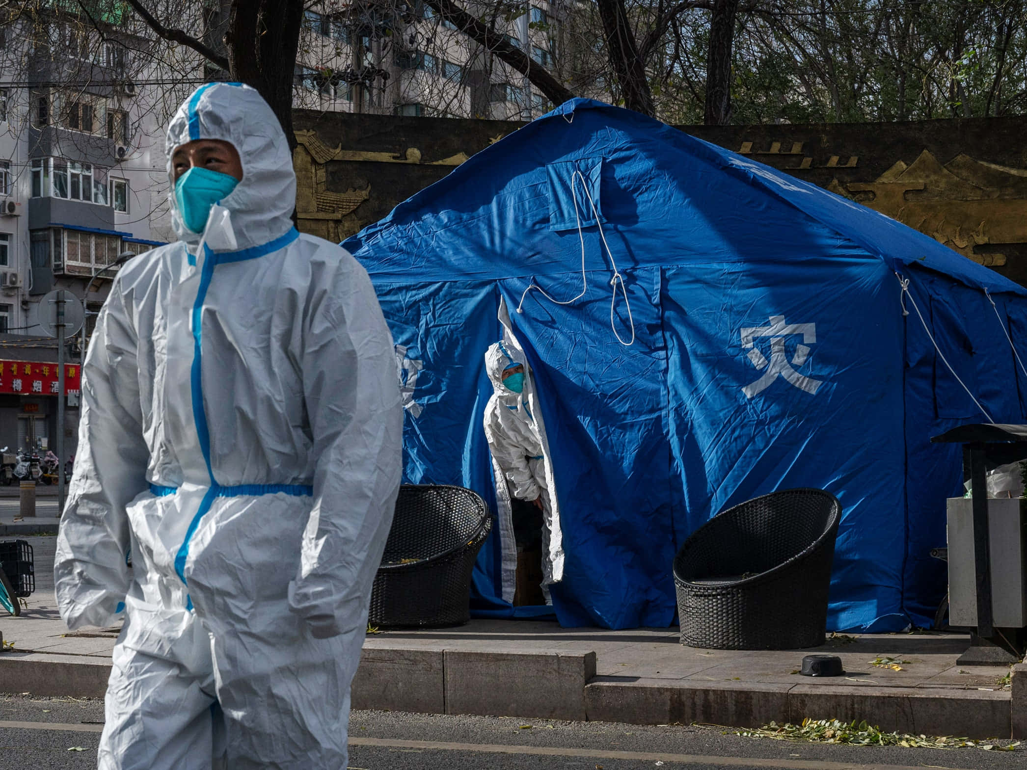 中國 桌布 壁紙 — 一名穿著白色防護服的醫護人員在都市環境中走過一個標有中文字的藍色應急帳篷。