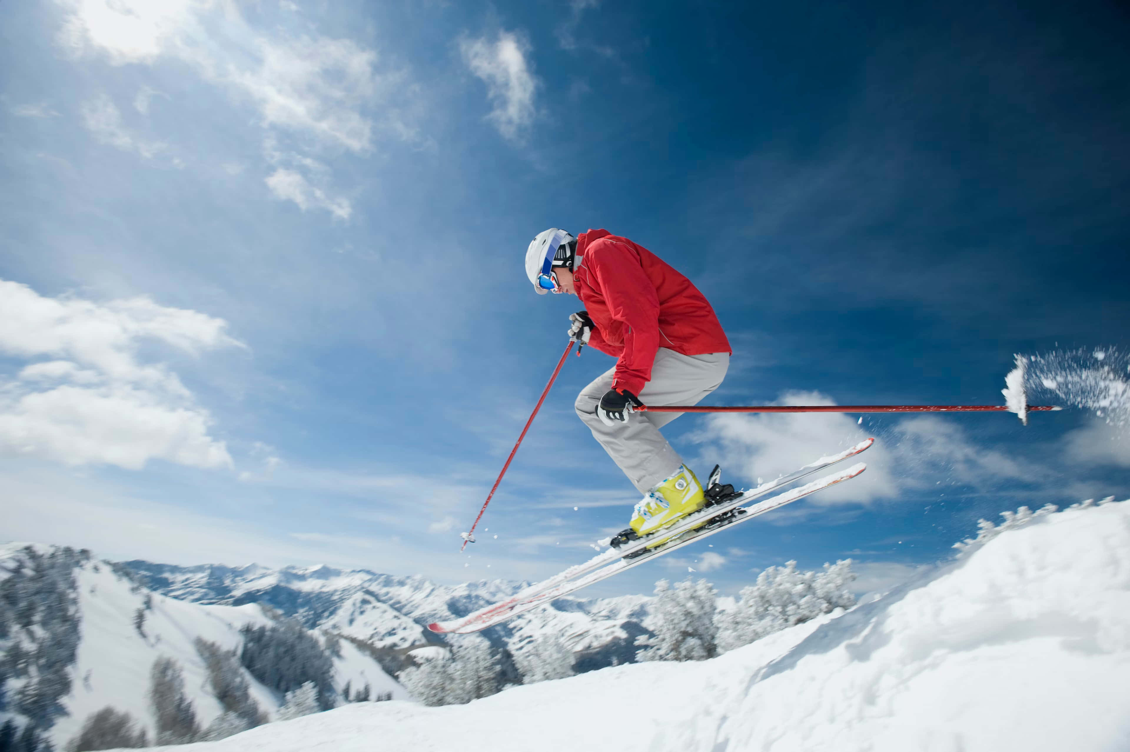 ski 桌布 壁紙 — 一名穿著紅色外套的滑雪者在明亮的藍天白雲下，飛越白雪皚皚的山坡。