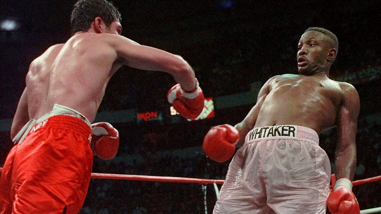 Boxing 桌布 壁紙 — 一名穿著紅色短褲的拳擊手在明亮的場館燈光下，於激烈的擂台比賽中向身穿白色短褲的對手揮拳。