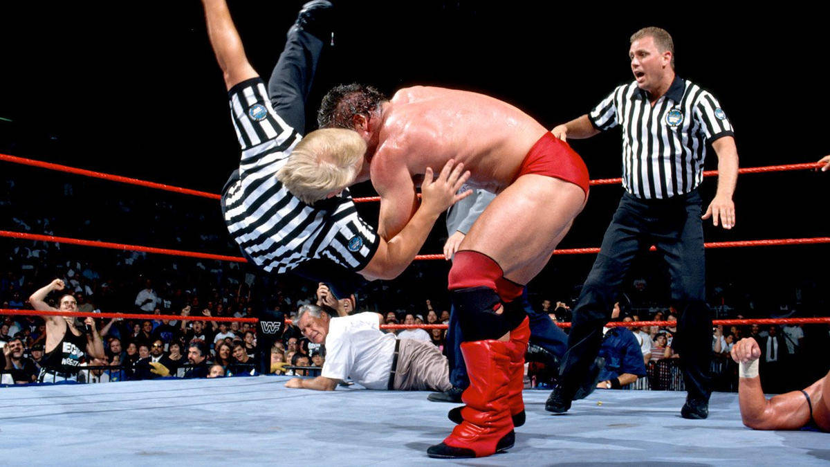 WWE 桌布 壁紙 — 一名穿著紅色緊身褲的摔角手在混亂的職業摔角比賽中將裁判舉到半空，背景有另一名摔角手和人群。