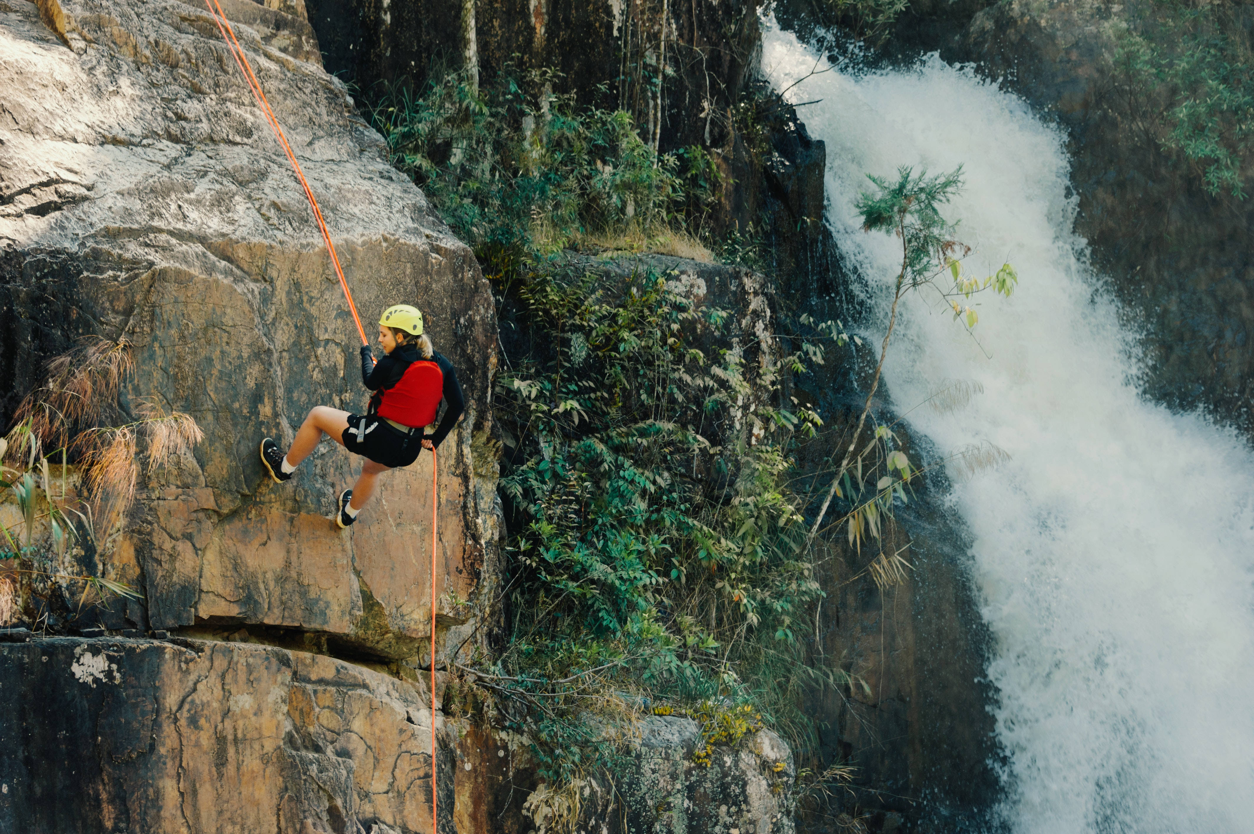 Climbing 桌布 壁紙 — 一名穿著紅色裝備的攀岩者正在陡峭的懸崖上繩降，旁邊是咆哮的瀑布，周圍環繞著茂密的綠色植物。