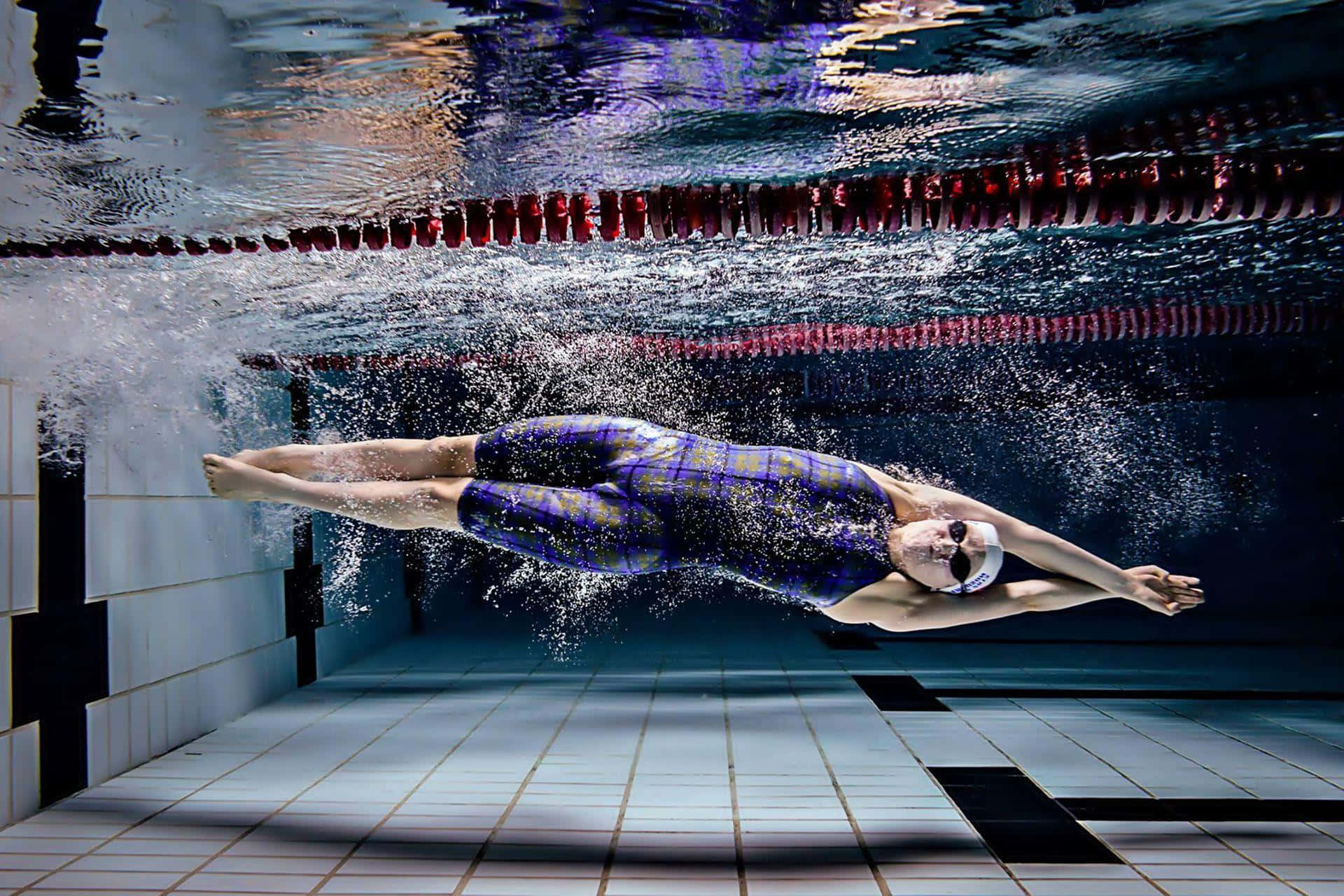 Swimming 桌布 壁紙 — 一名穿著紫色圖案泳衣的泳手在設有紅色泳道分隔和瓷磚牆的泳池中潛水滑行。