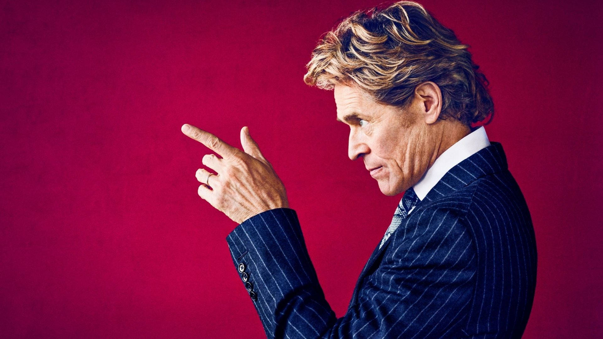 Willem Dafoe 桌布 壁紙 — 一名穿著細條紋西裝的男子在鮮紅色背景下權威地指著。