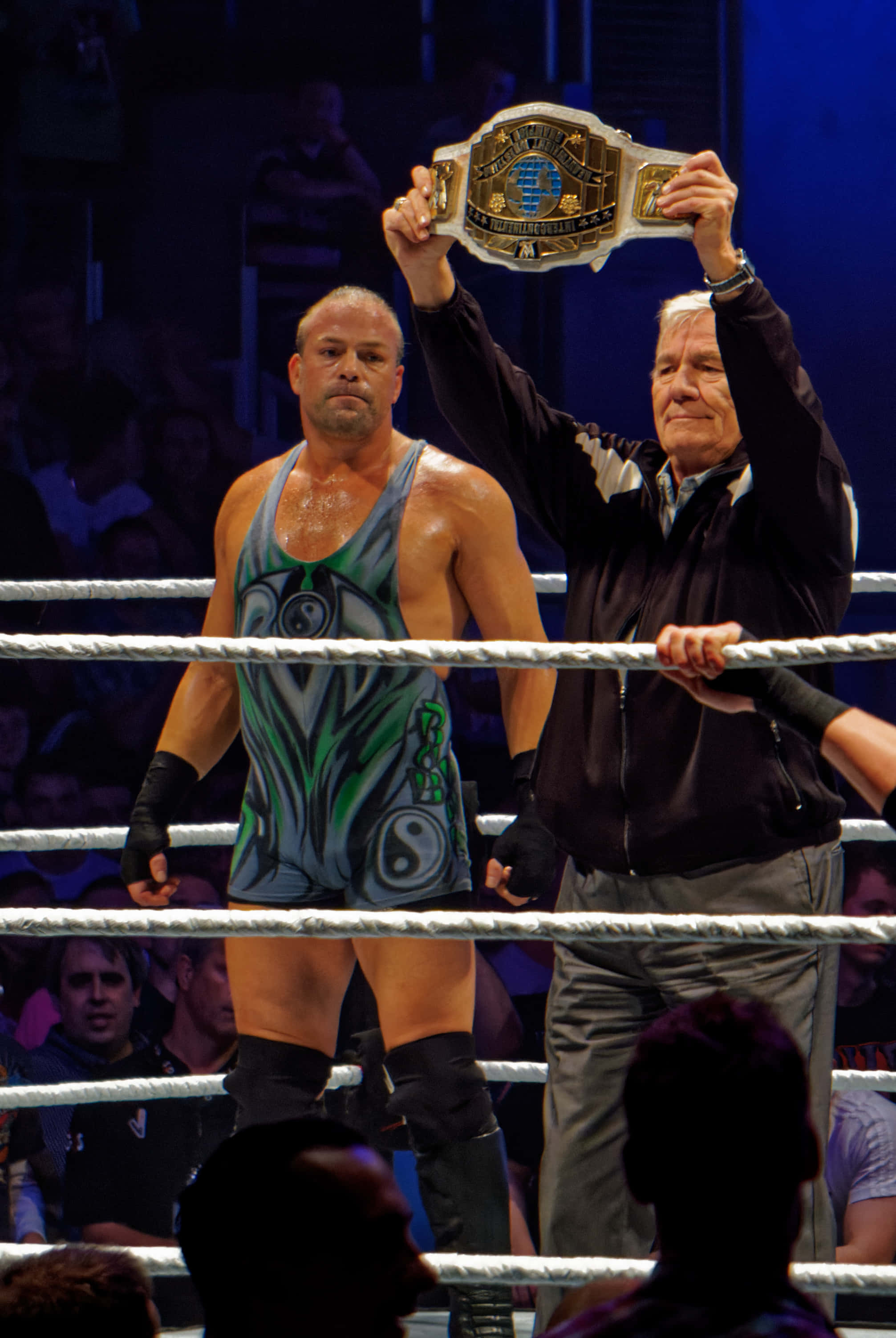 WWE 桌布 壁紙 — 一名穿著綠色和藍色緊身衣的摔角手站在擂台上，一位年長男子為觀眾將冠軍腰帶高舉過頭。