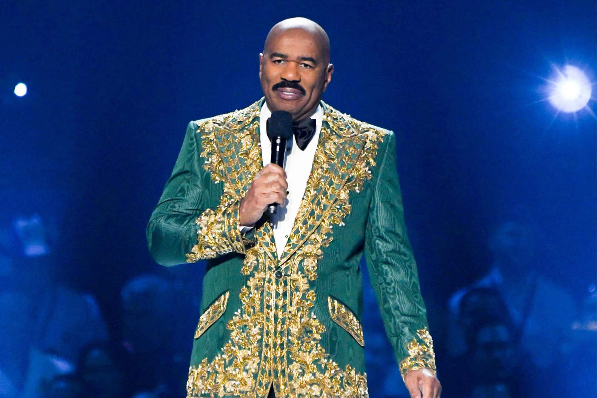 steve harvey 桌布 壁紙 — 一名穿著綠色和金色刺繡西裝的男子在明亮的舞台燈光下拿著麥克風。
