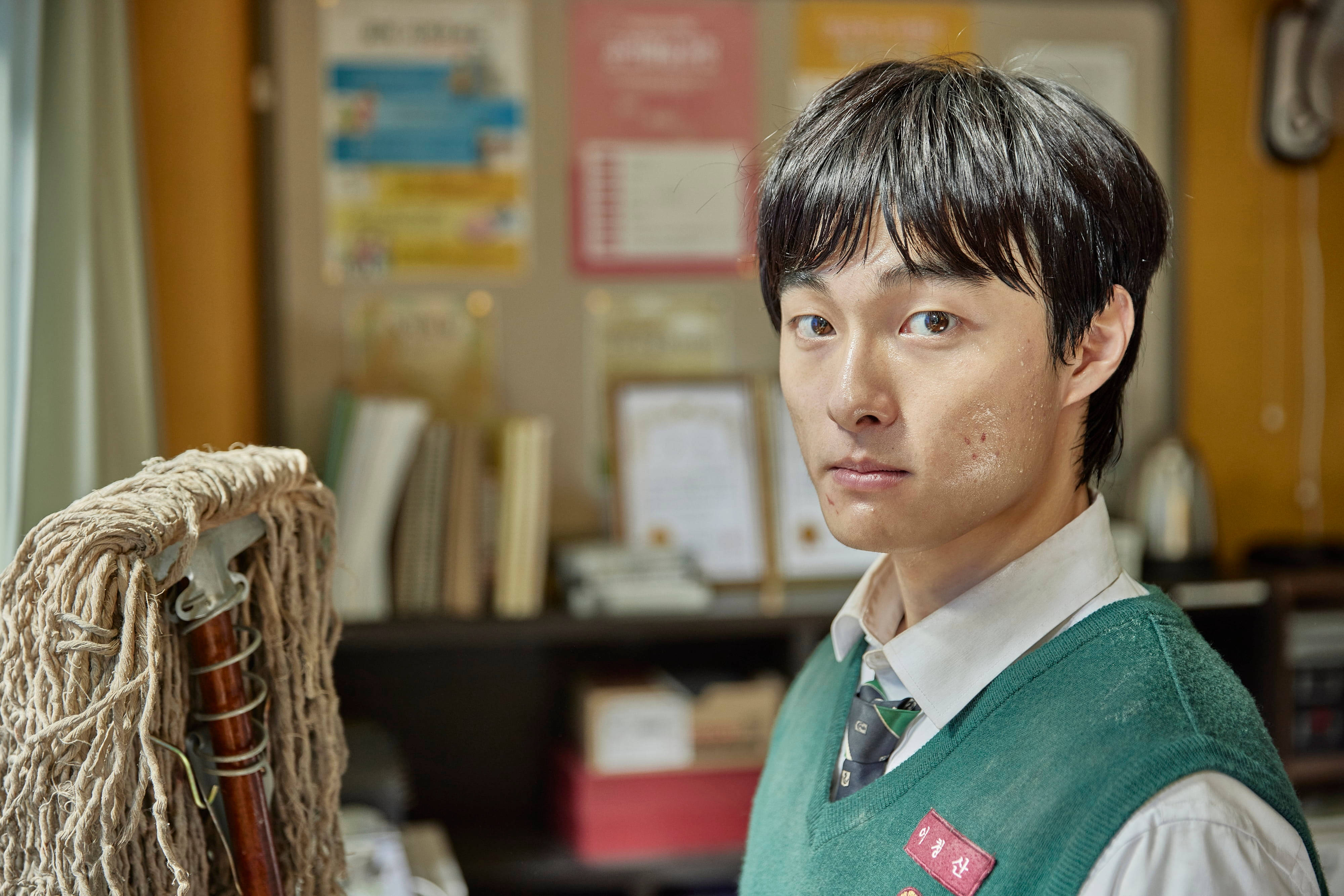 殭屍動圖 桌布 壁紙 — 一名穿著綠色背心和領帶的年輕男子，站在雜亂的辦公室裡，拿著拖把，表情嚴肅。
