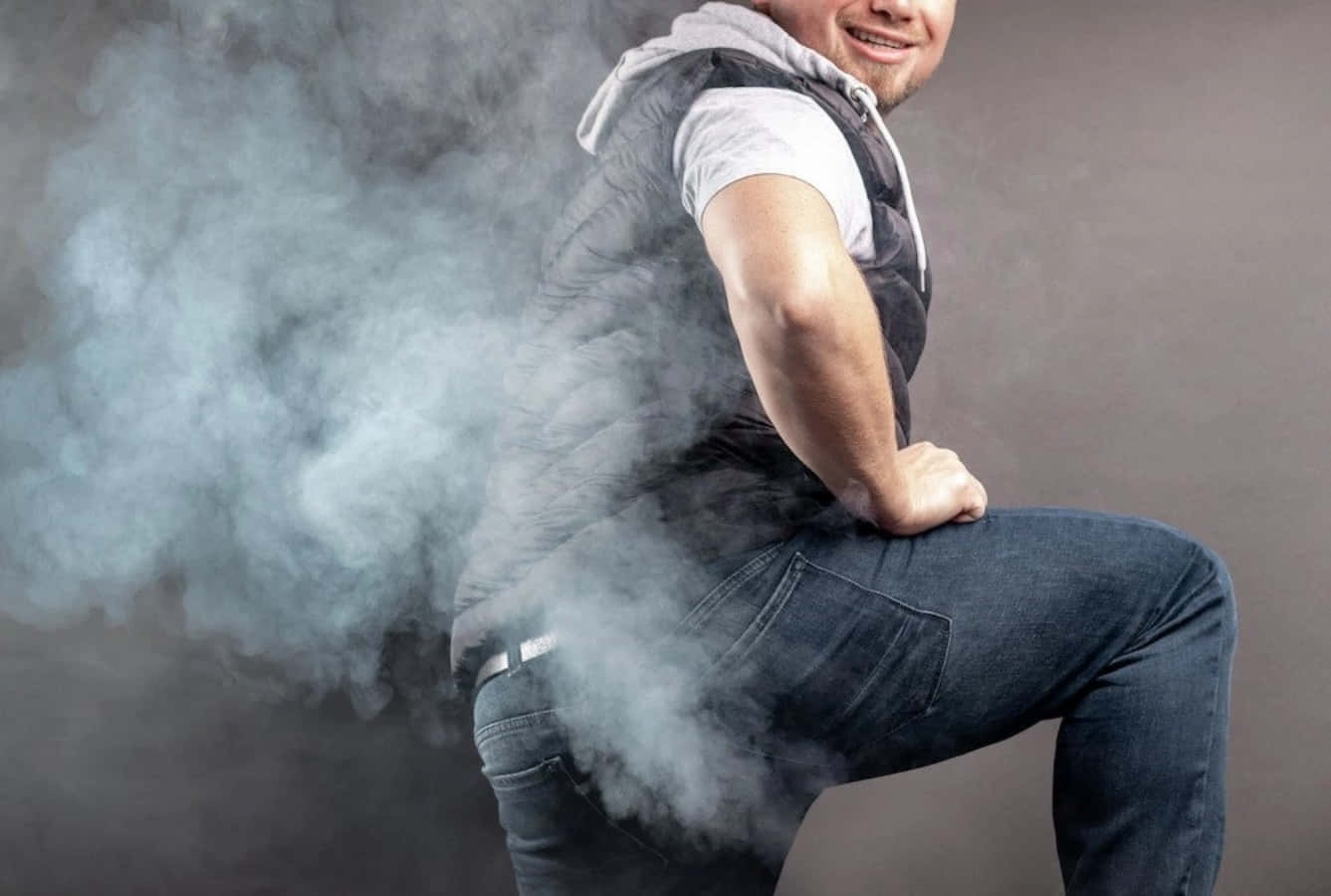 Fart 桌布 壁紙 — 一名穿著背心和牛仔褲的男子蹲下，背部冒出滾滾濃煙，背景為灰色攝影棚。