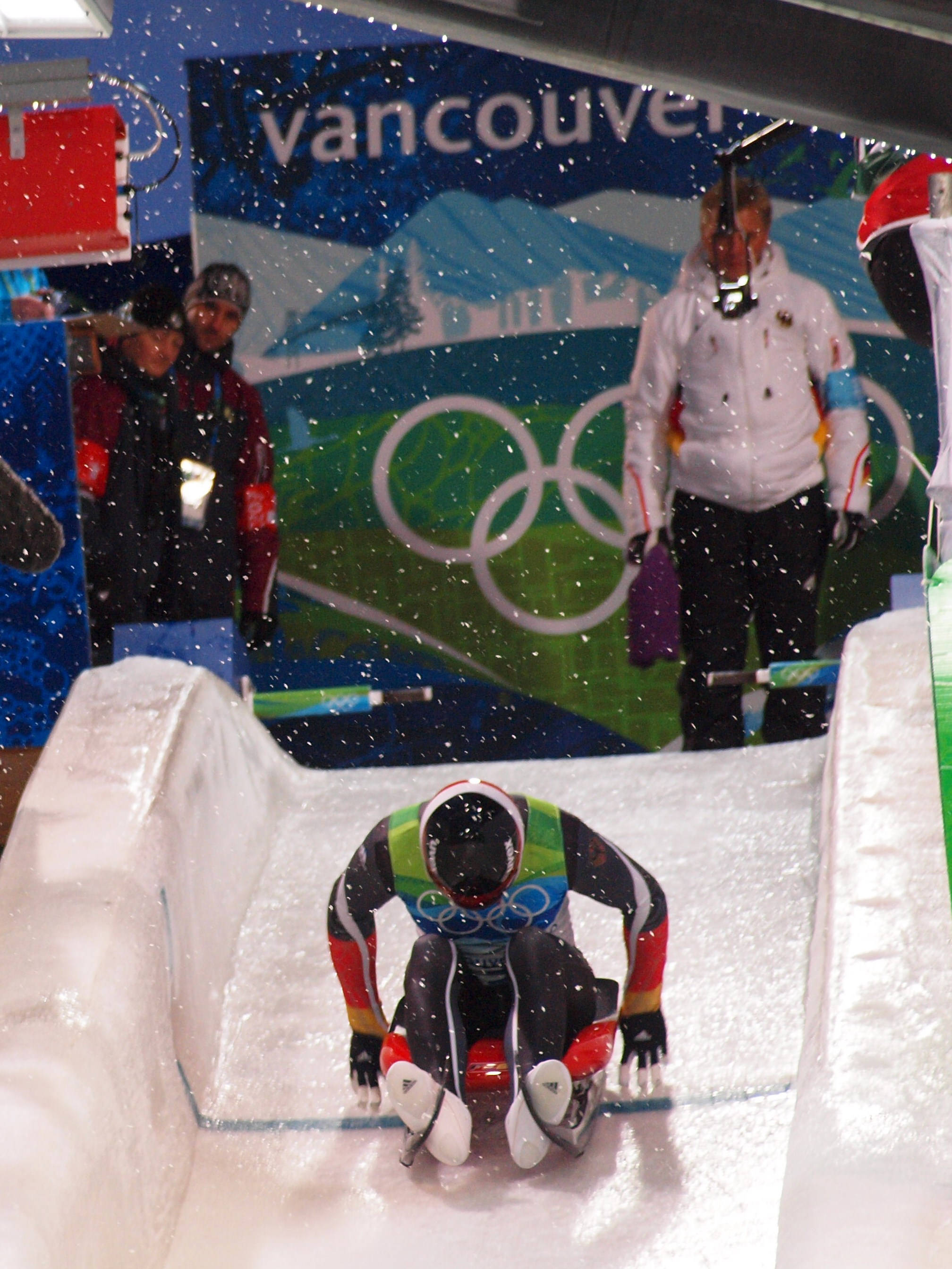 Olympic 桌布 壁紙 — 一名穿著色彩繽紛裝備的雪橇運動員在冰道起點蹲下，雪花飄落在奧運官員和溫哥華背景周圍。