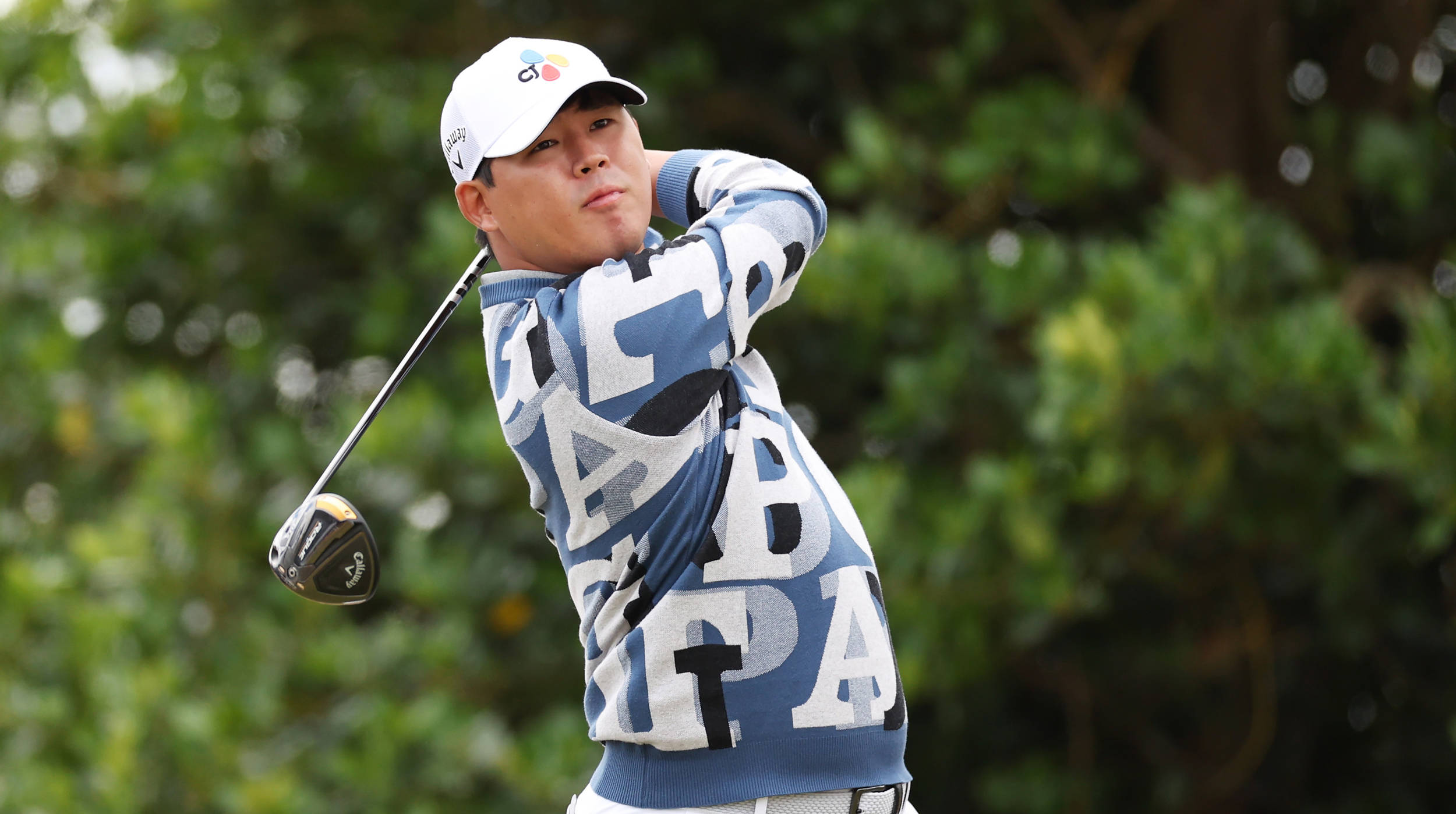 Athlete 桌布 壁紙 — 一名穿著藍色圖案毛衣的高爾夫球手，在模糊的綠色樹葉背景下，專注地揮桿。