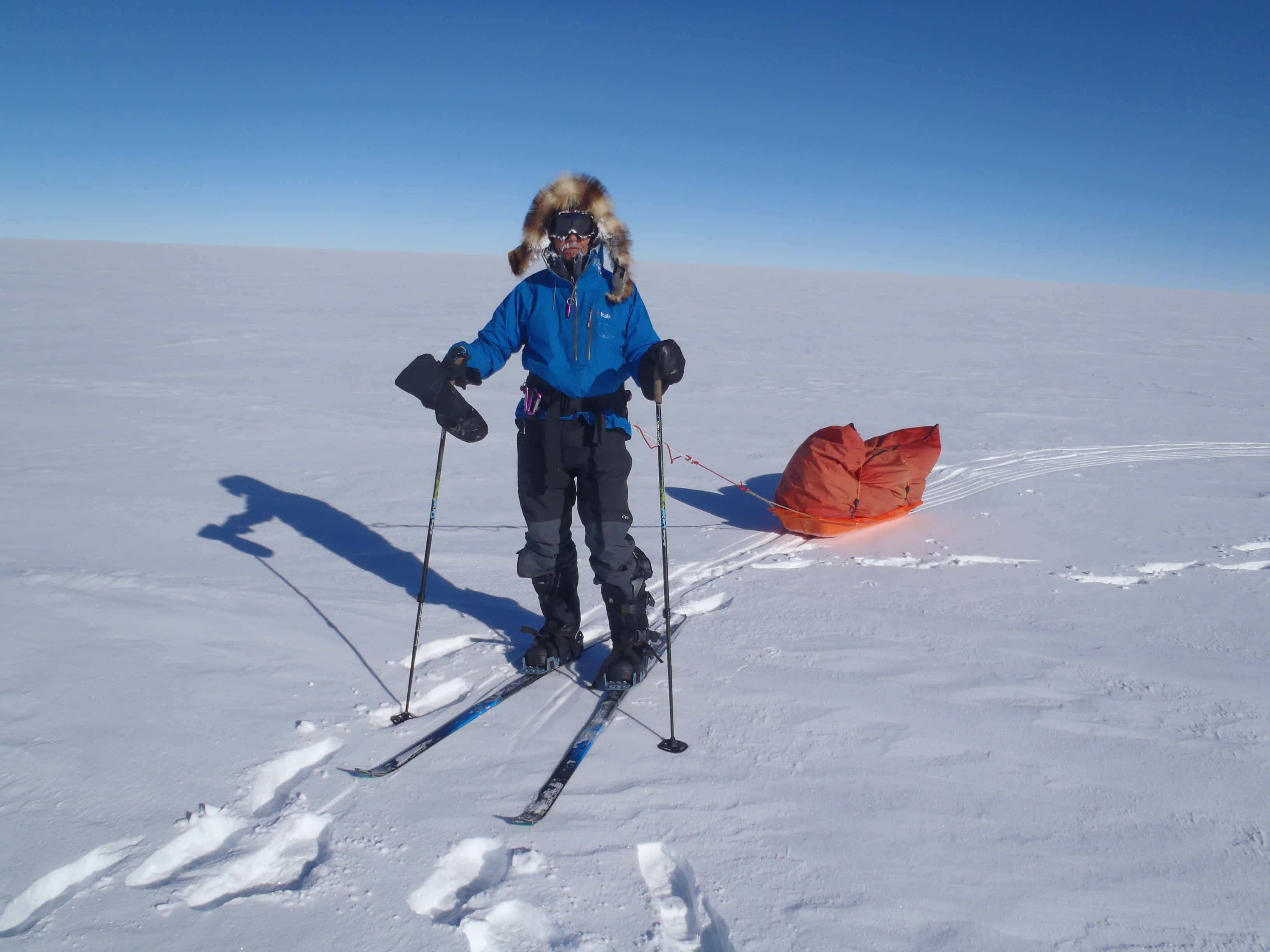 South Pole 桌布 壁紙 — 一名穿著藍色外套和毛邊兜帽的滑雪者站在雪地上，附近有滑雪杖和一個橙色袋子，天空清澈湛藍。