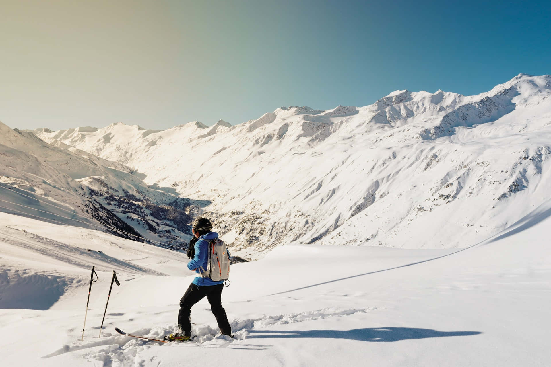 ski 桌布 壁紙 — 一名穿著藍色外套的滑雪者，在晴朗藍天和陽光普照的山峰下，穿越廣闊的雪山山谷。