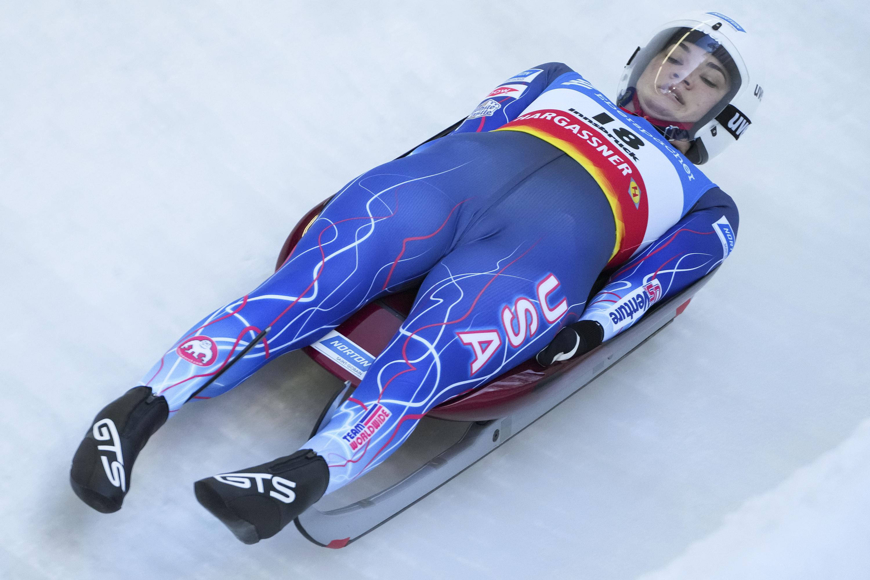 Austria 桌布 壁紙 — 一名穿著藍色服裝、帶有紅色和白色點綴的雪橇運動員，戴著頭盔和護目鏡在冰道上飛馳。