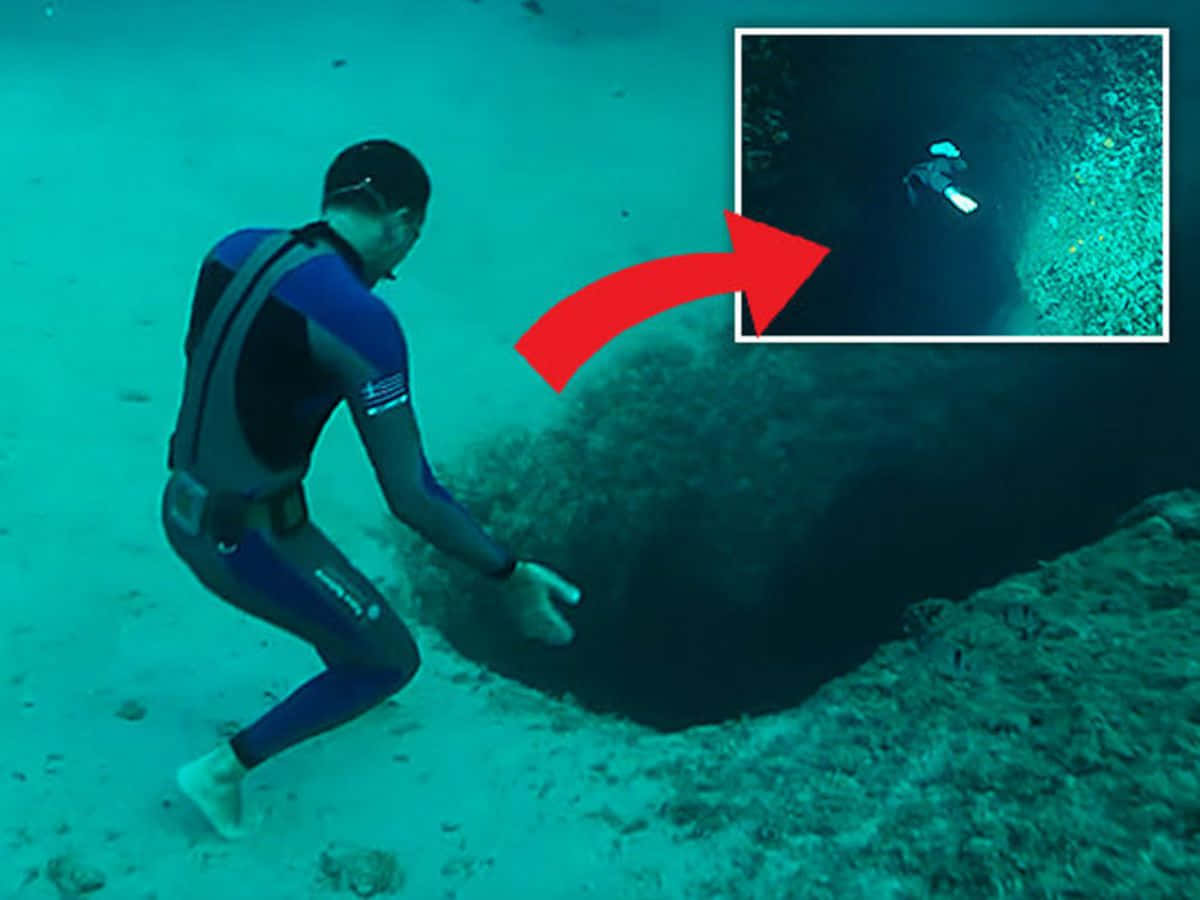 Scary Underwater 桌布 壁紙 — 一名穿著藍色潛水衣的潛水員跪在一個黑暗的水下洞穴入口旁，周圍是碧綠的海水和沙質海床。