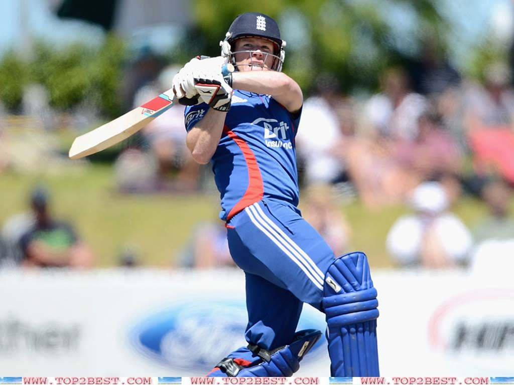 eoin morgan 桌布 壁紙 — 一名穿著藍色裝備的板球擊球手在明亮的日光下揮動球棒，背景有模糊的觀眾。