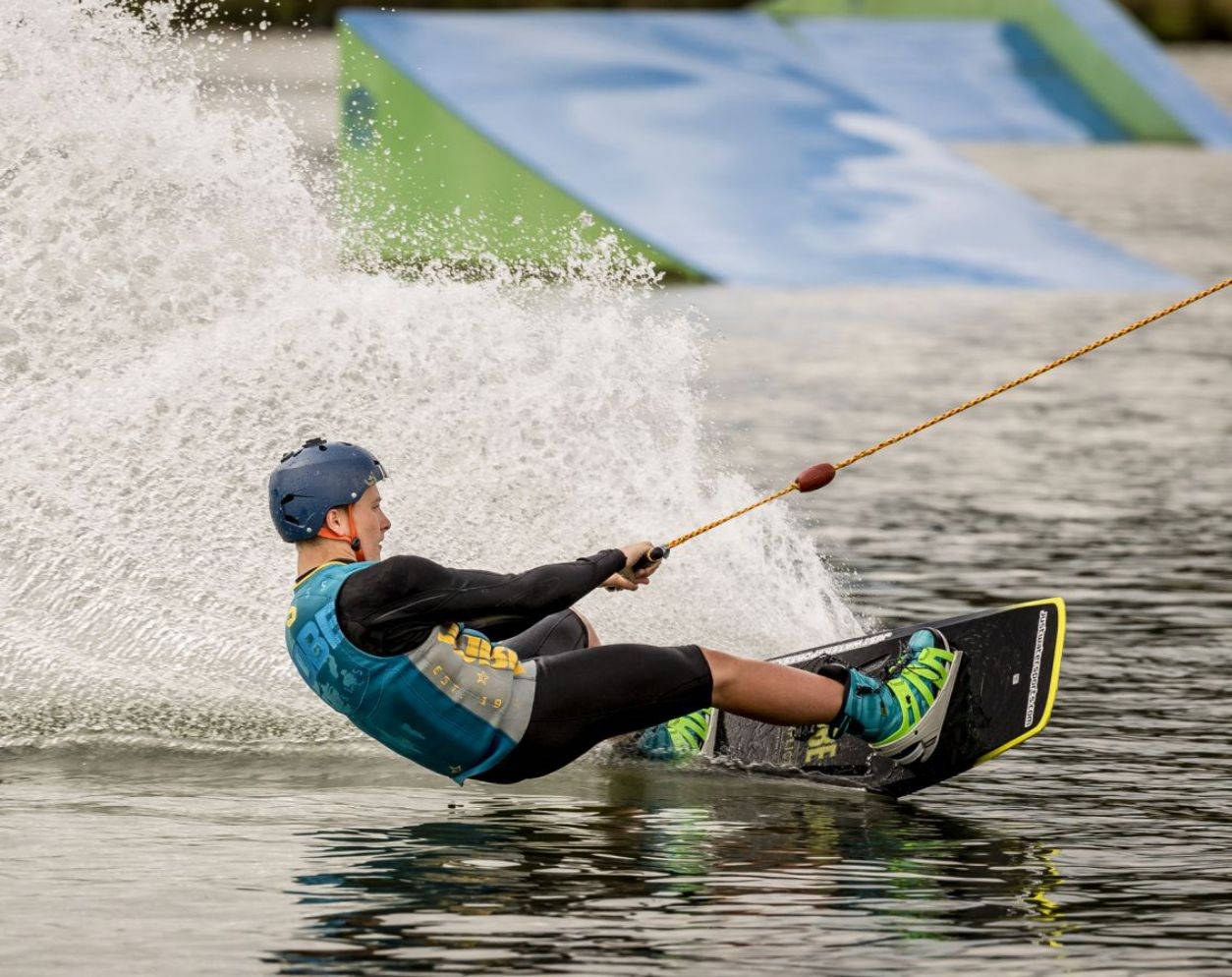 surfing 桌布 壁紙 — 一名穿著藍色頭盔和背心的滑水運動員在湖上劃過身後濺起的水花，背景是色彩繽紛的跳台。