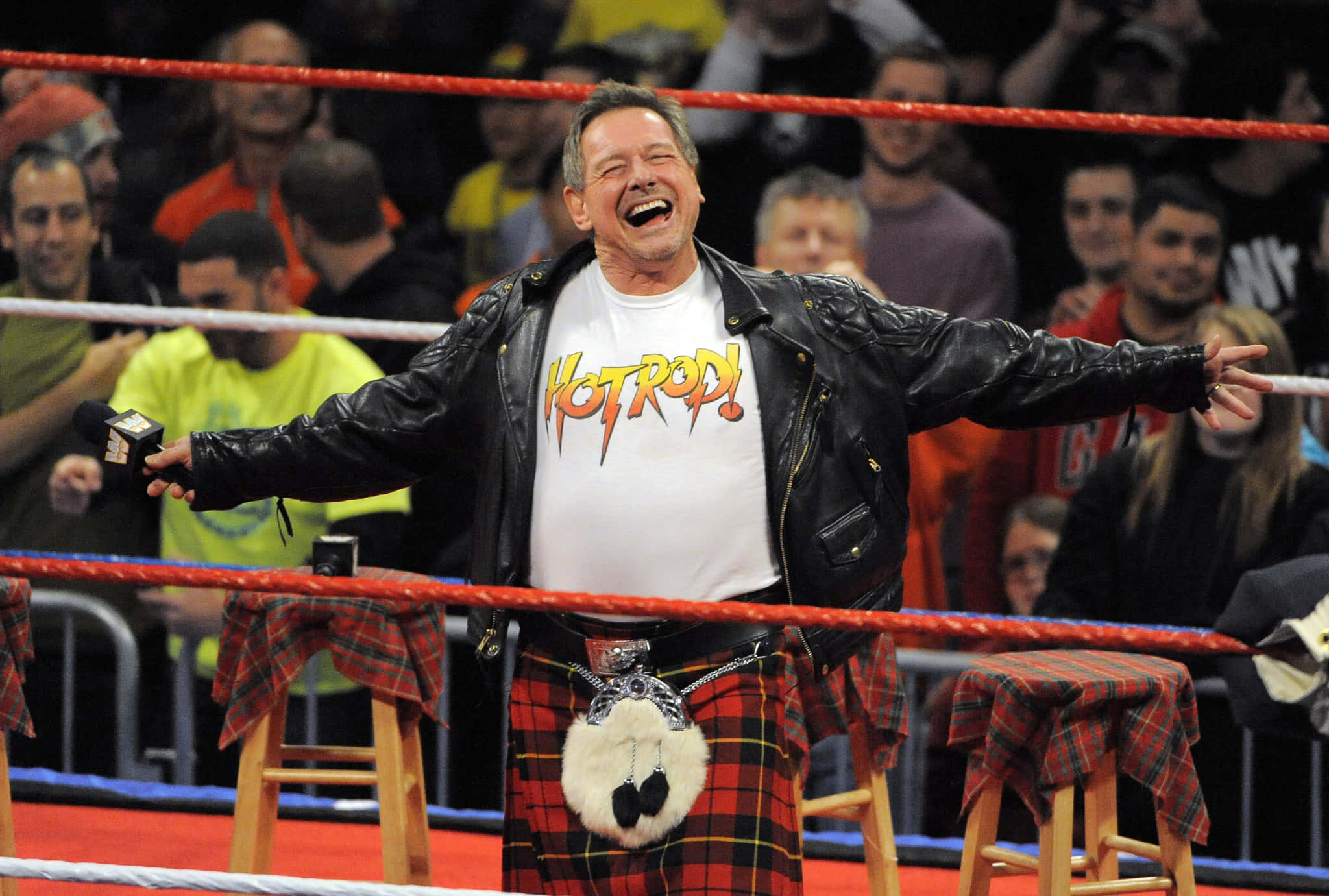 WWE 桌布 壁紙 — 一名穿著蘇格蘭裙和皮夾克的摔角手在擂台上開懷大笑，身後有粉絲為他歡呼。